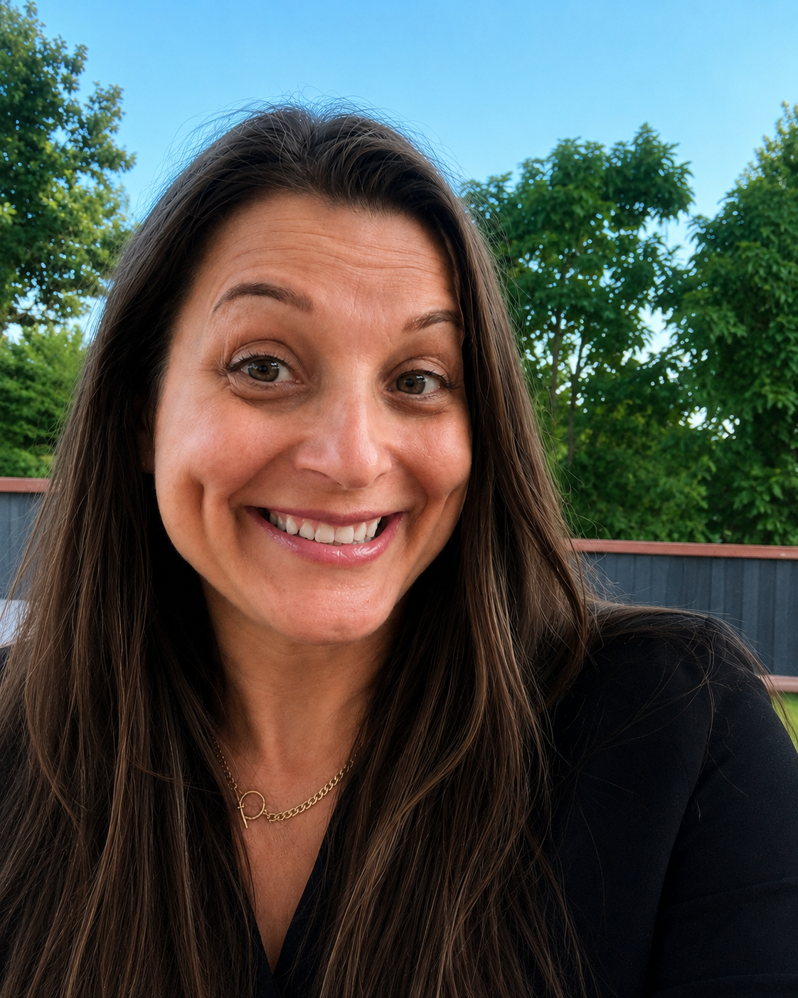 Smiling woman outdoors in a black top, with trees and a blue sky in the background