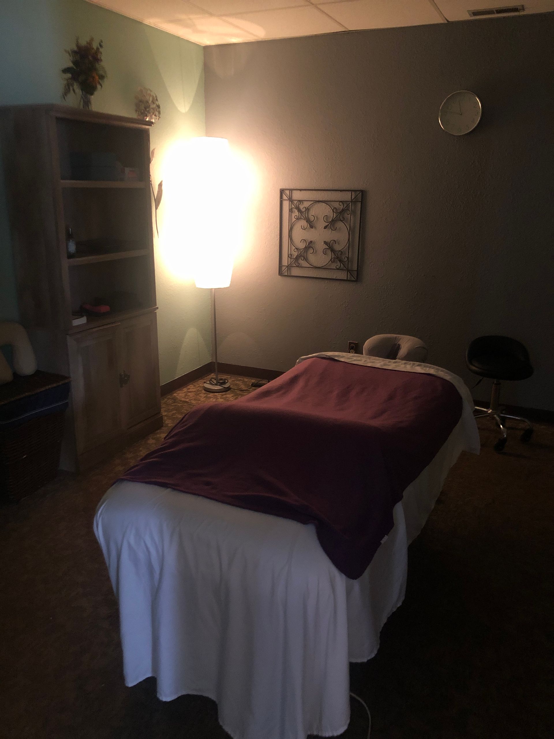 Dim massage room with a treatment table, burgundy blanket, wall art, and a bright lamp.