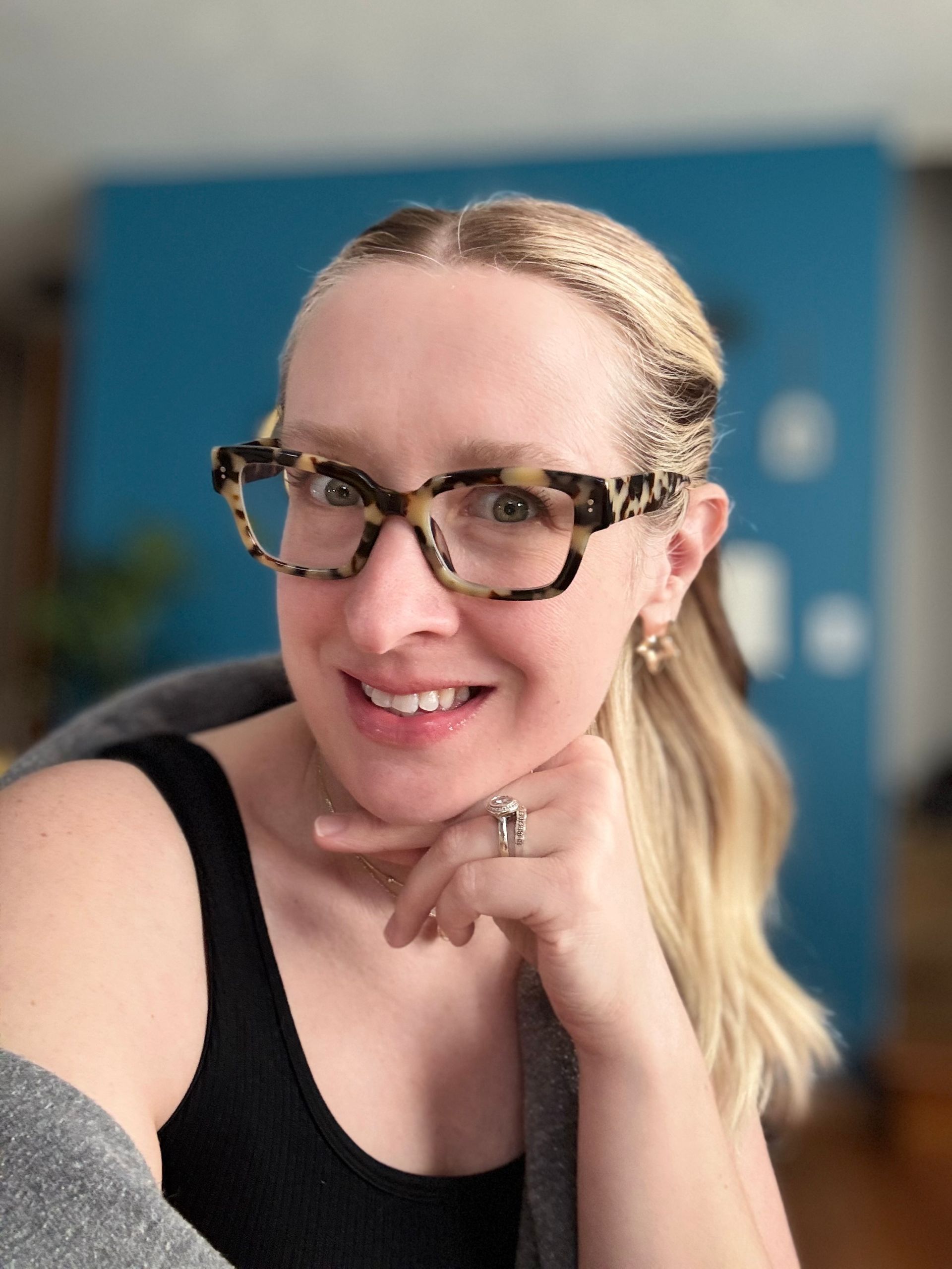 Smiling person in tortoiseshell glasses, seated indoors with a blue wall background.
