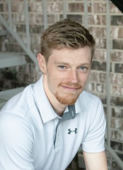 Man in a white polo shirt smiling in front of a brick wall