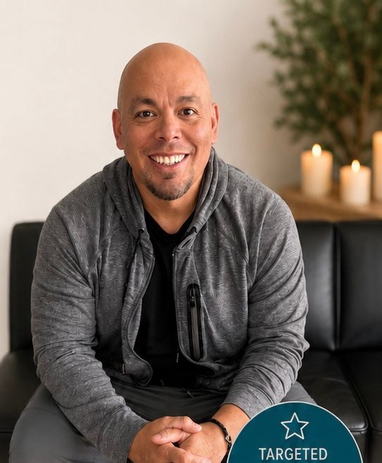 Smiling bald man in a gray cardigan sits on a black couch in a softly lit room with candles.