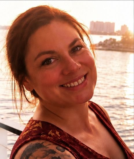 Smiling woman on a boat at sunset, with waterfront buildings in the background.