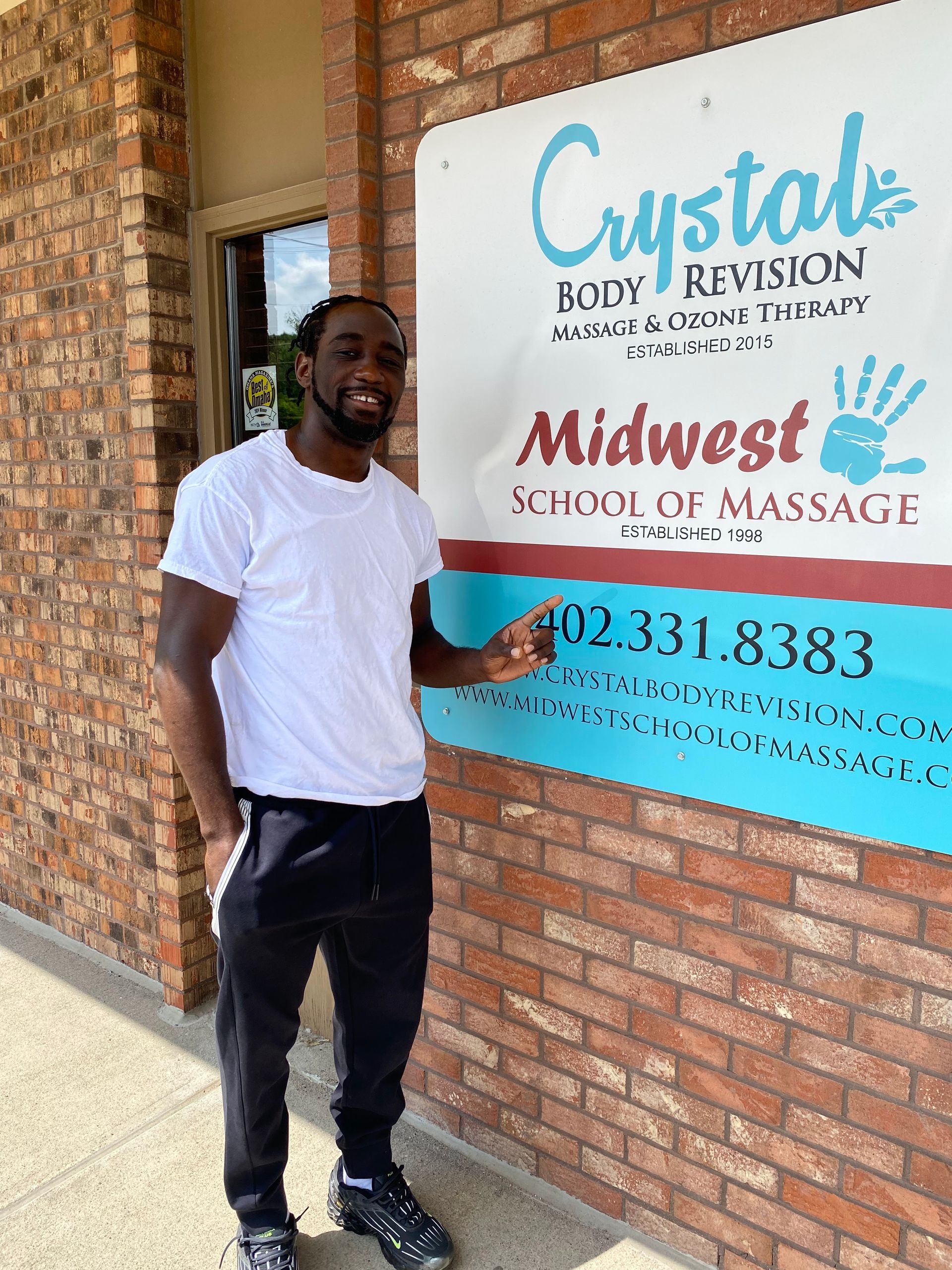 Man standing beside a brick wall sign for Crystal’s and Midwest School of Massage, smiling and pointing at it
