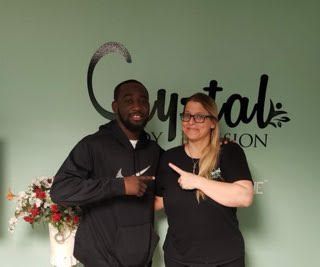 Two people posing and pointing at each other in front of a green salon wall sign.