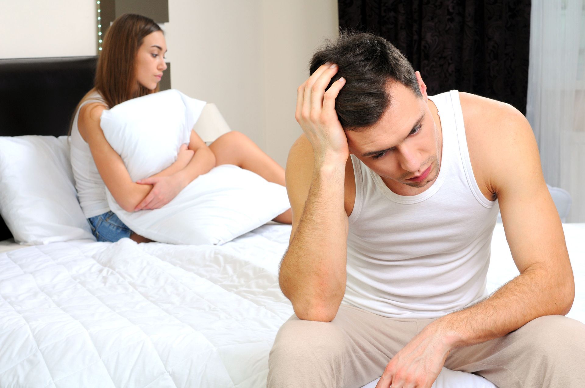 Man sits on bed, head in hand, looking distressed. Woman sits behind, clutching pillow, looking away.