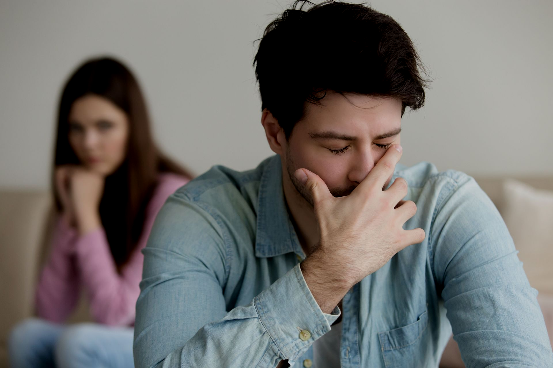 Man with hand on face looks distressed, woman in background appears pensive, on a couch.