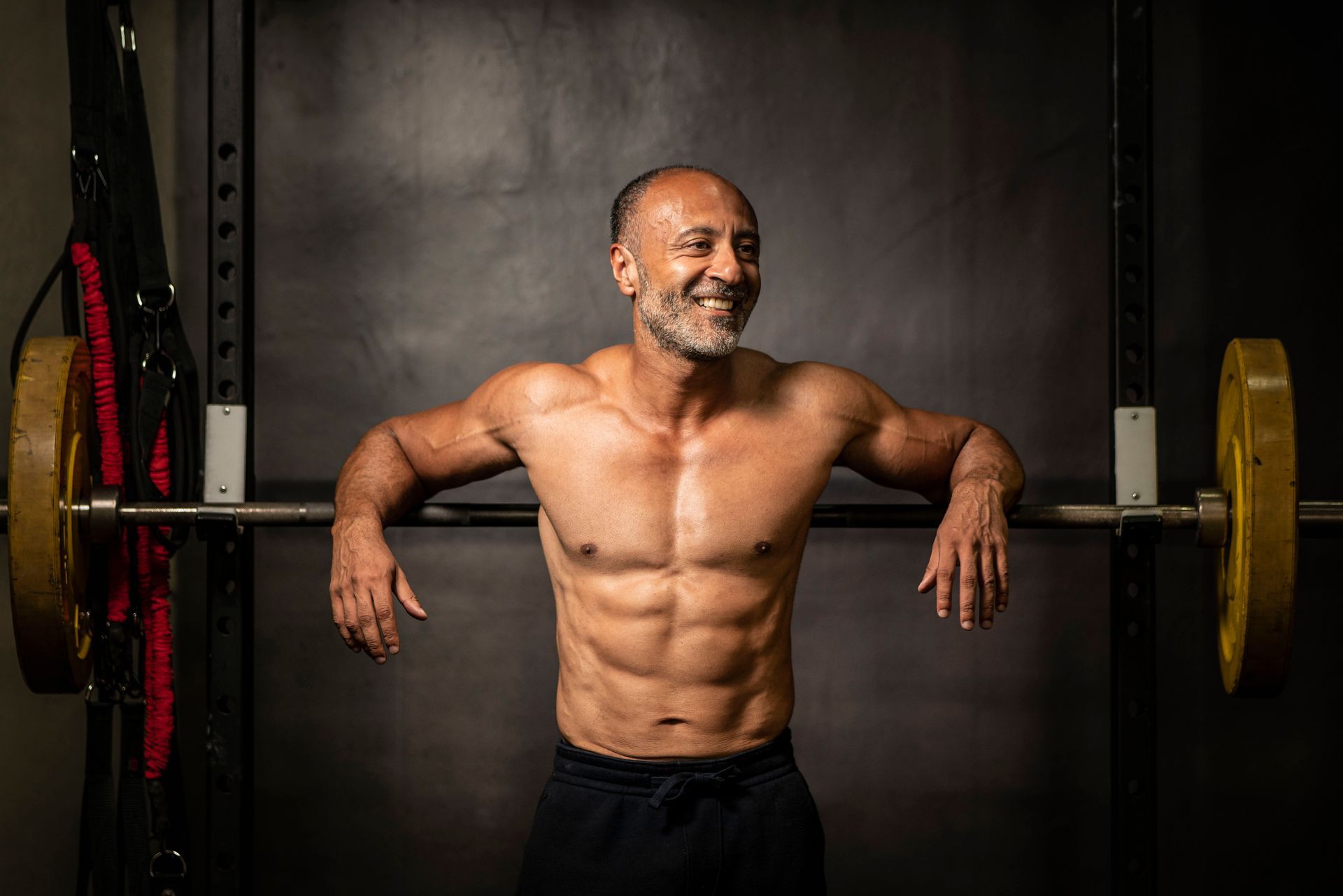 Muscular man with abs smiles, leaning on a barbell rack in a gym.