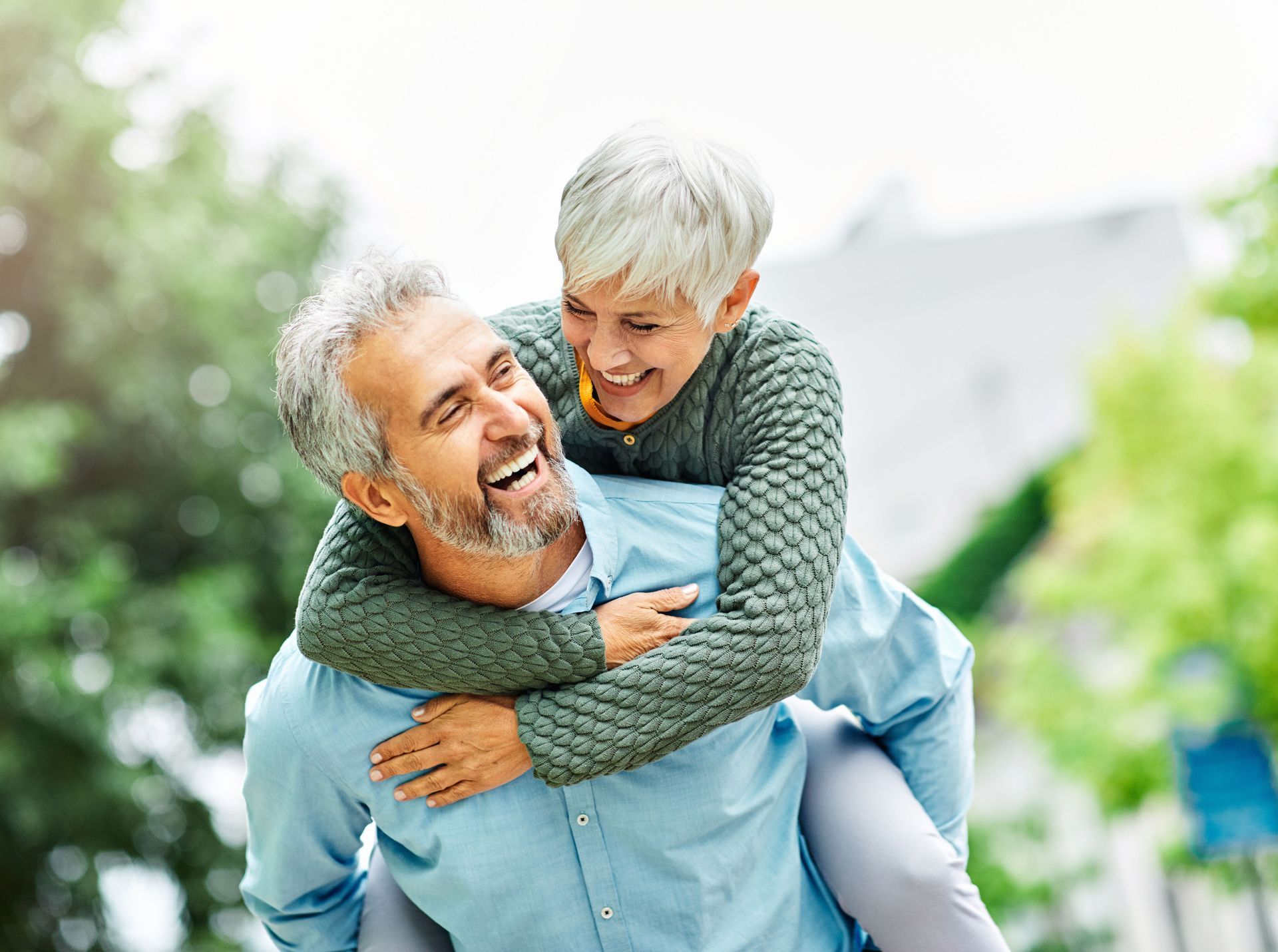 Older man gives a woman a piggyback ride, both laughing outdoors.
