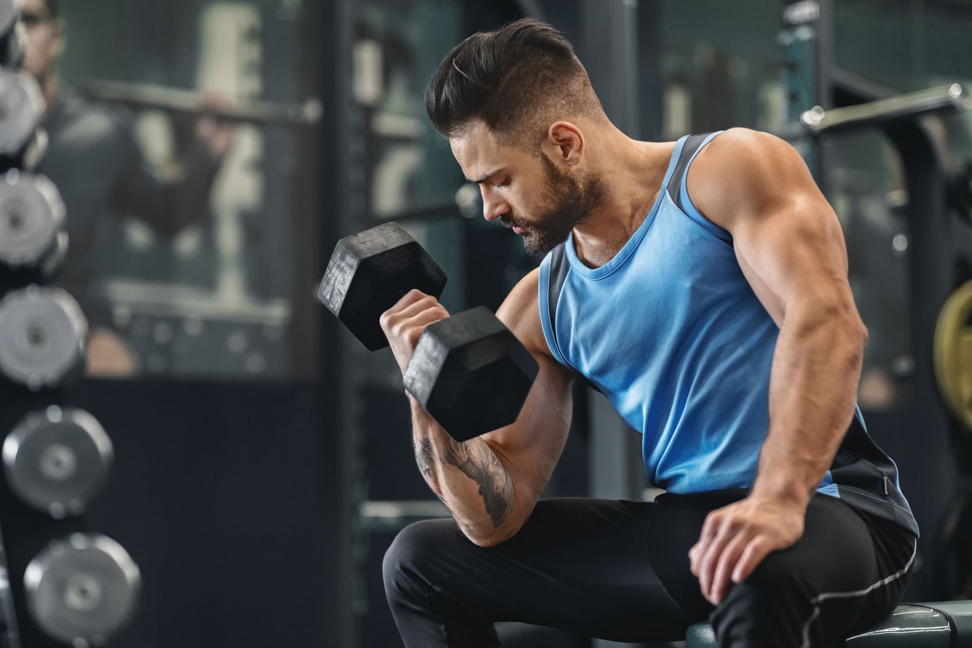 Man in a gym curls dumbbell while seated, wearing blue tank top and black pants.