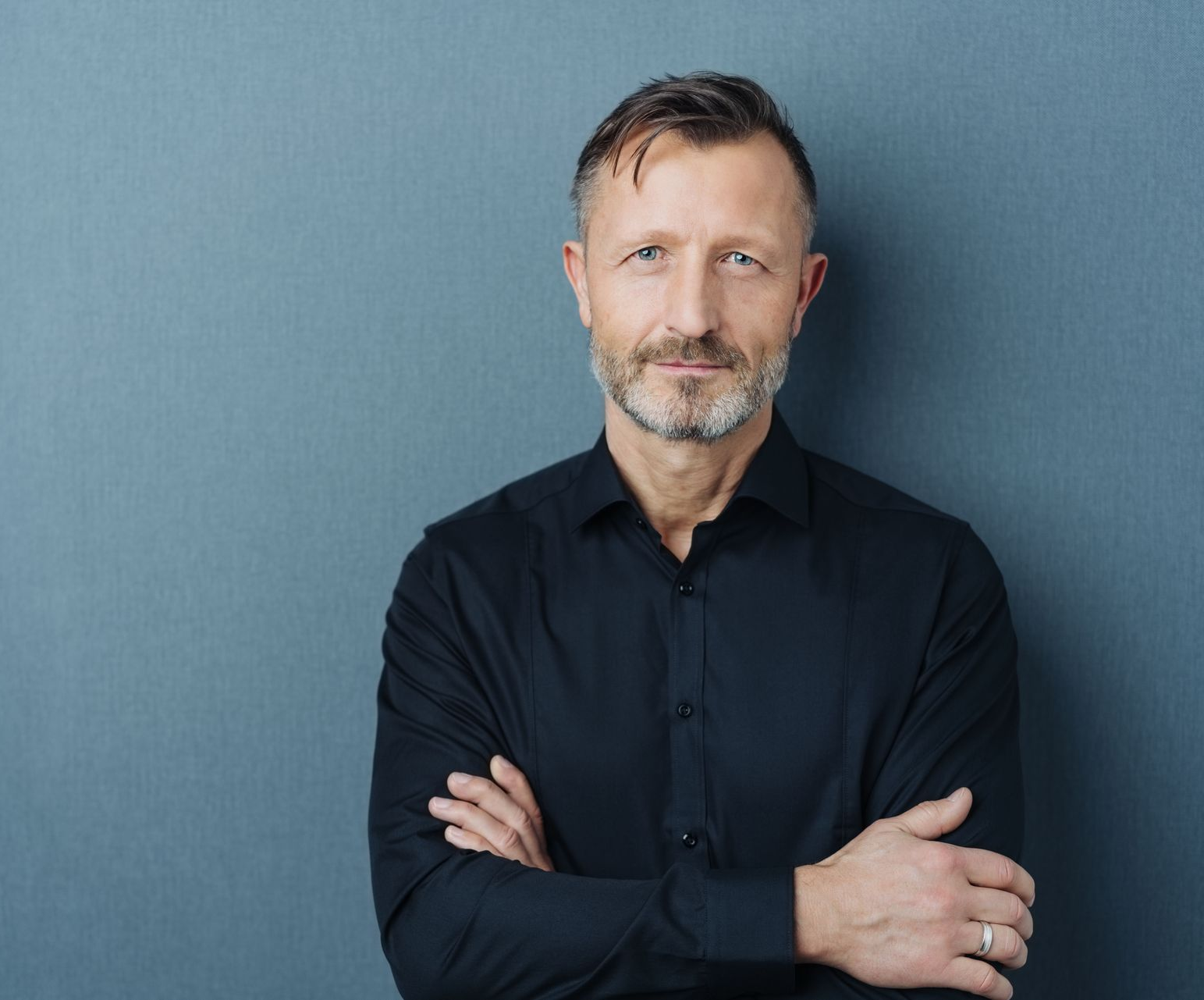 Man with crossed arms, graying hair and beard, wearing a black shirt, standing against a blue wall.