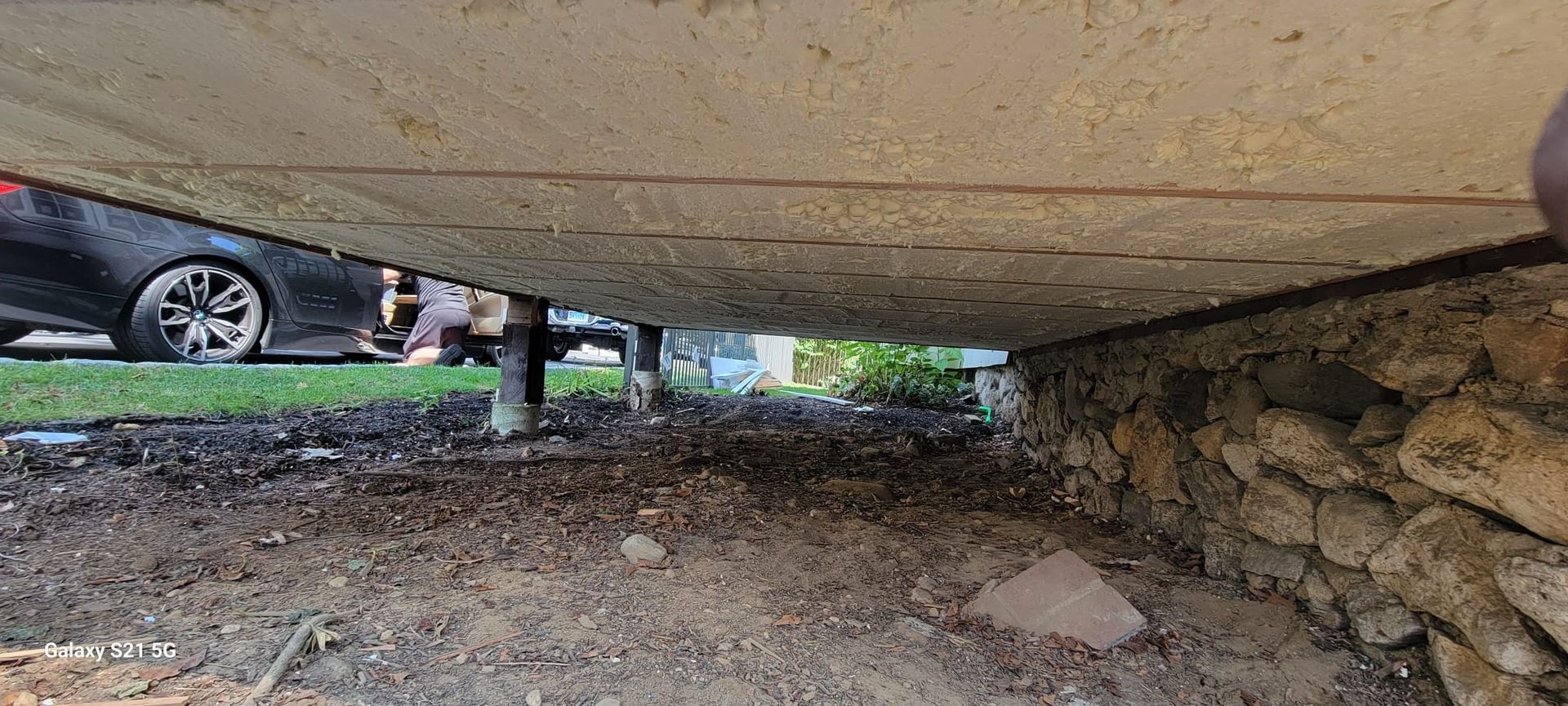 A car is parked under a concrete structure next to a stone wall.