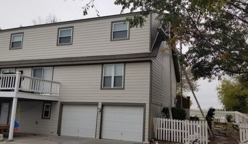 Three-story beige townhouse with garage doors, white fence, and a tree in front
