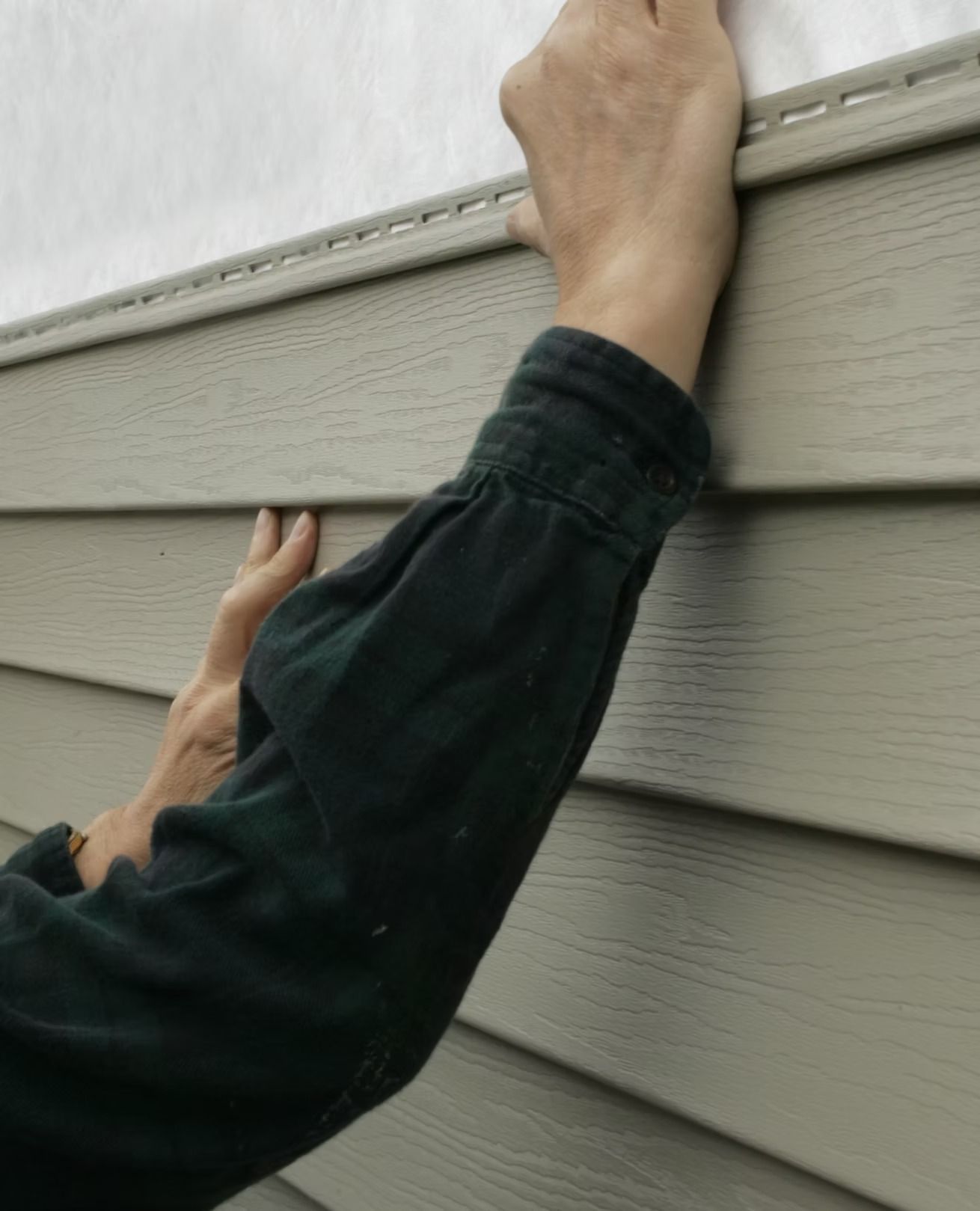 Person reaching up and gripping the top of a gray house siding wall
