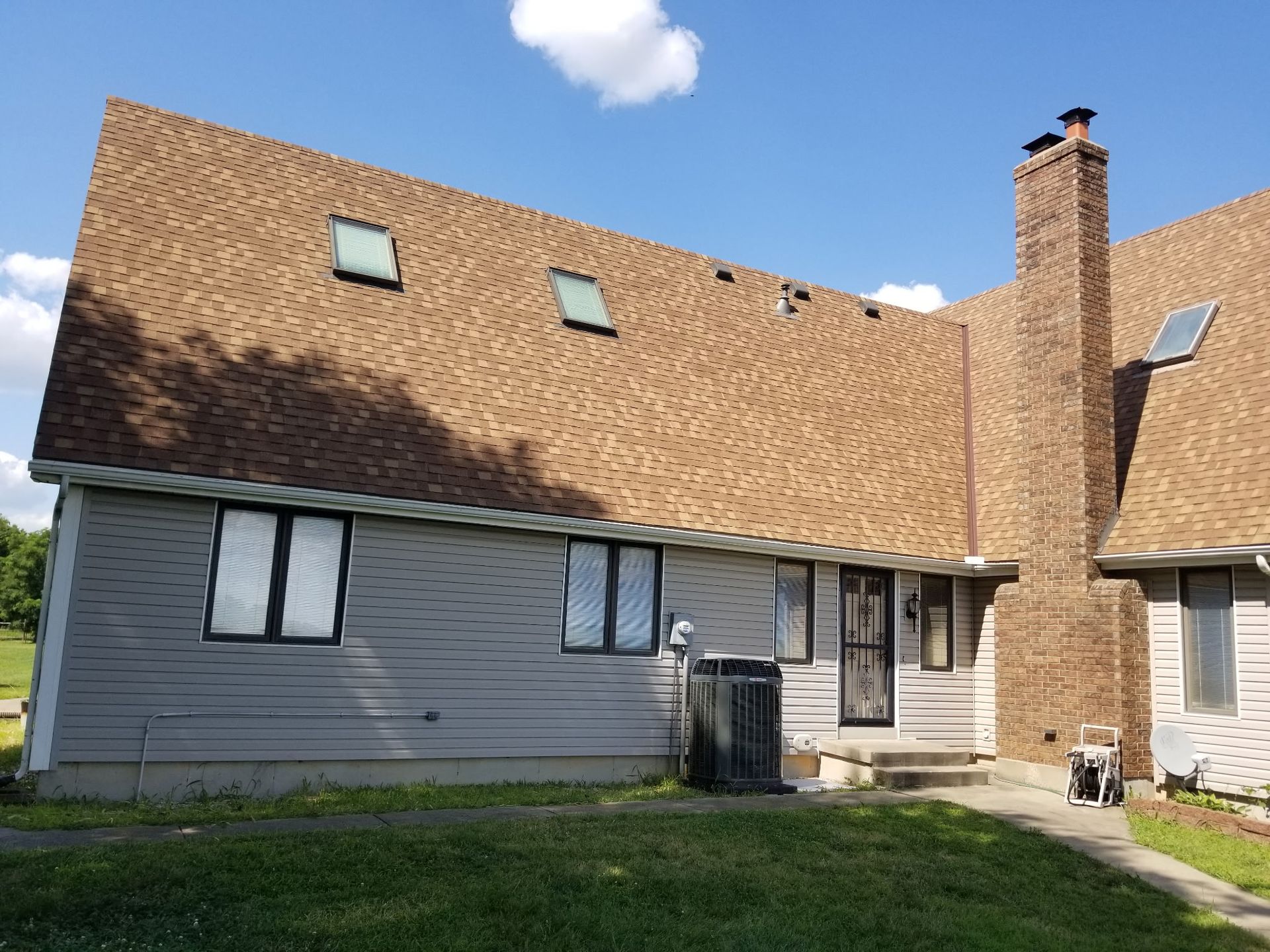 House with tan shingle roof, gray siding, brick chimney, and blue sky with clouds
