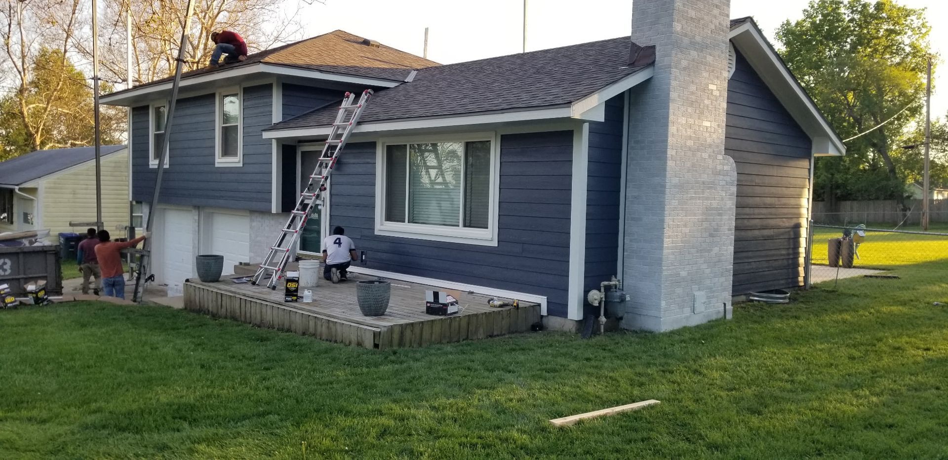 Blue house with stone chimney and patio in a grassy yard, with a person on the porch