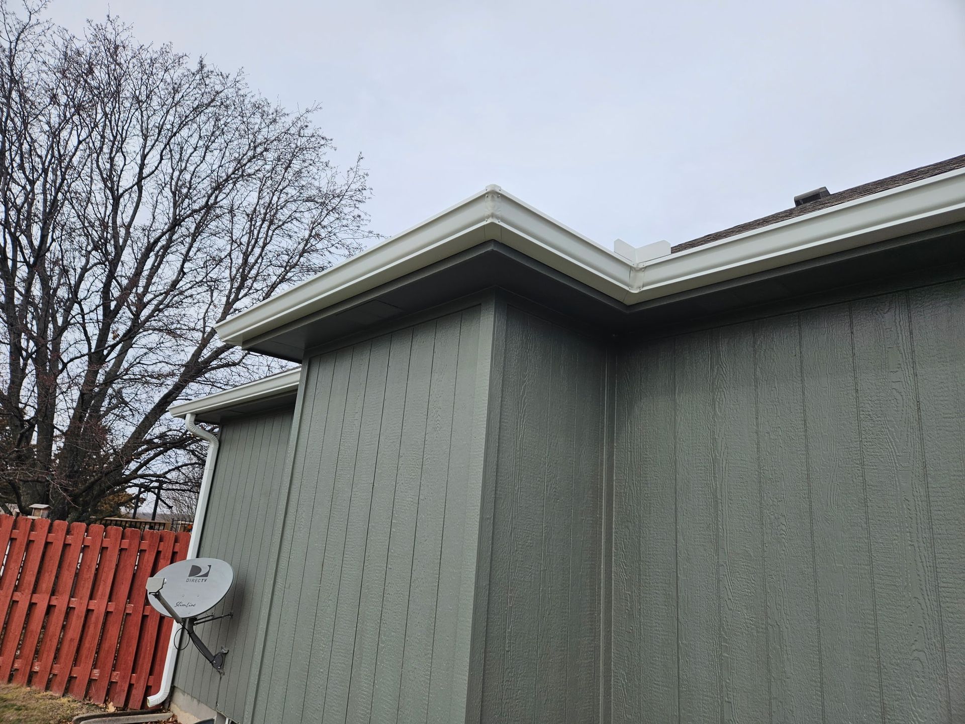 Gray house exterior with satellite dish, red fence, and bare trees under an overcast sky