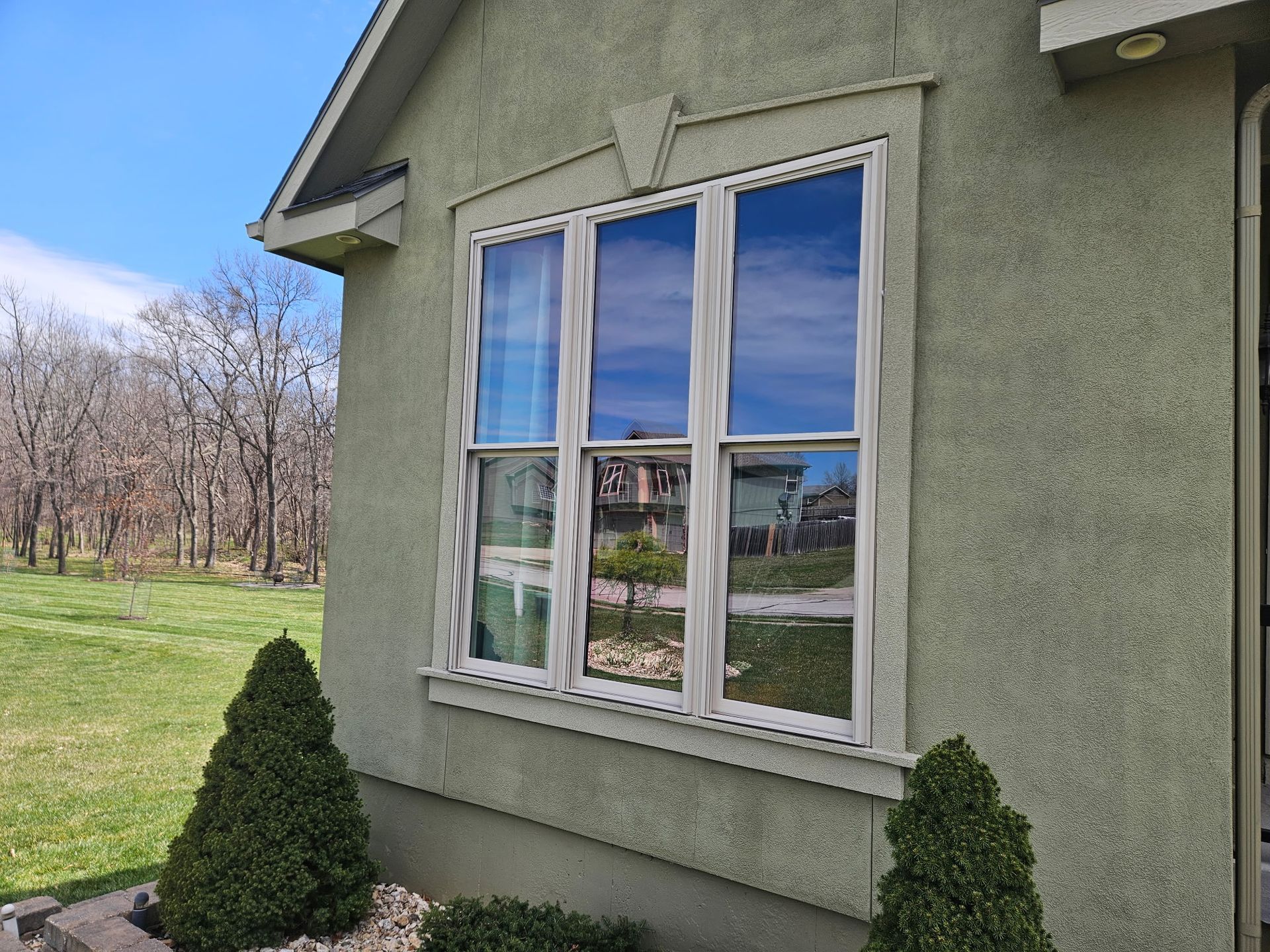 House exterior with a double window, green stucco wall, and small shrubs beside a grassy yard.