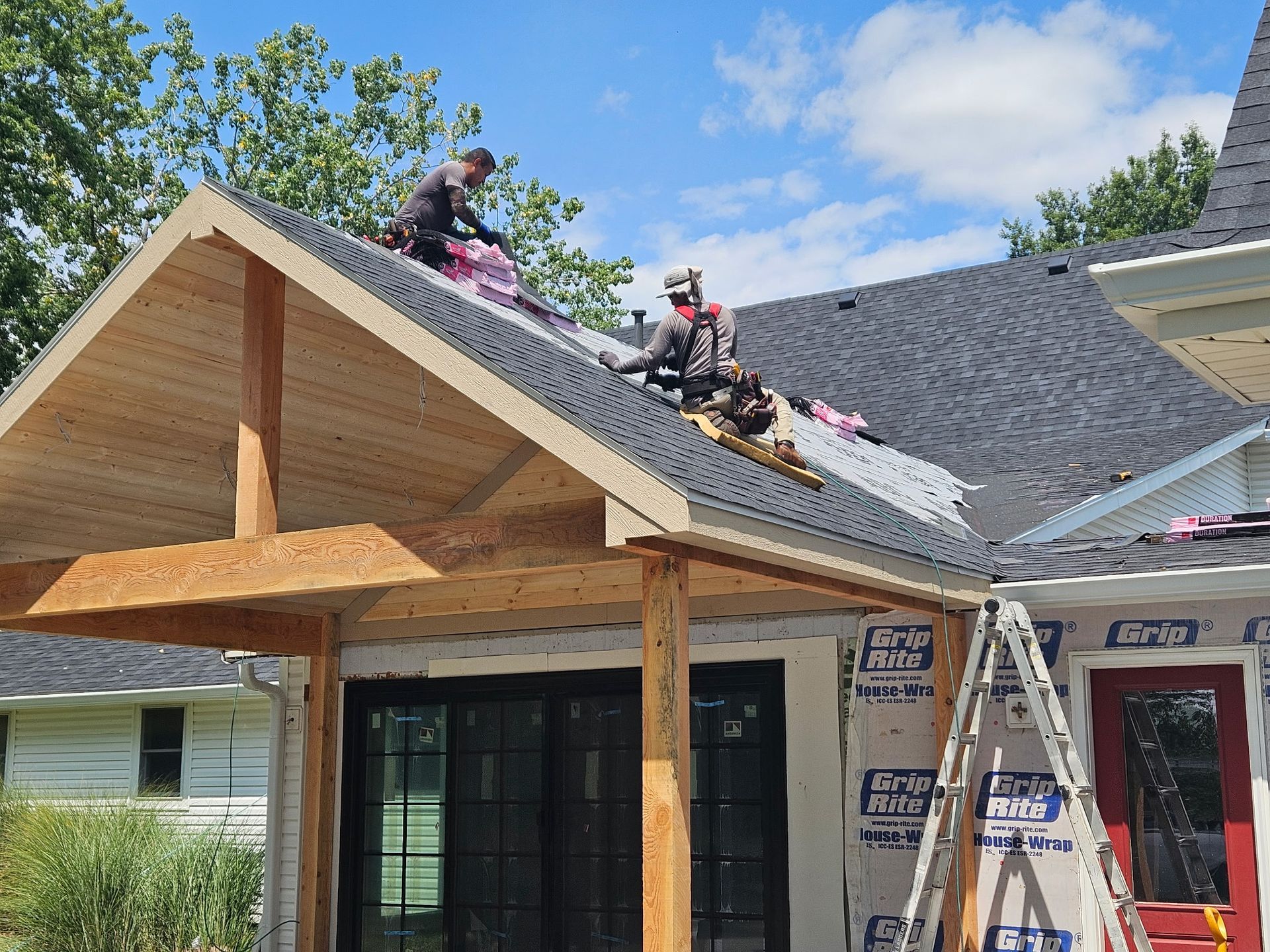 Workers on a house roof installing shingles on a porch addition under a blue sky