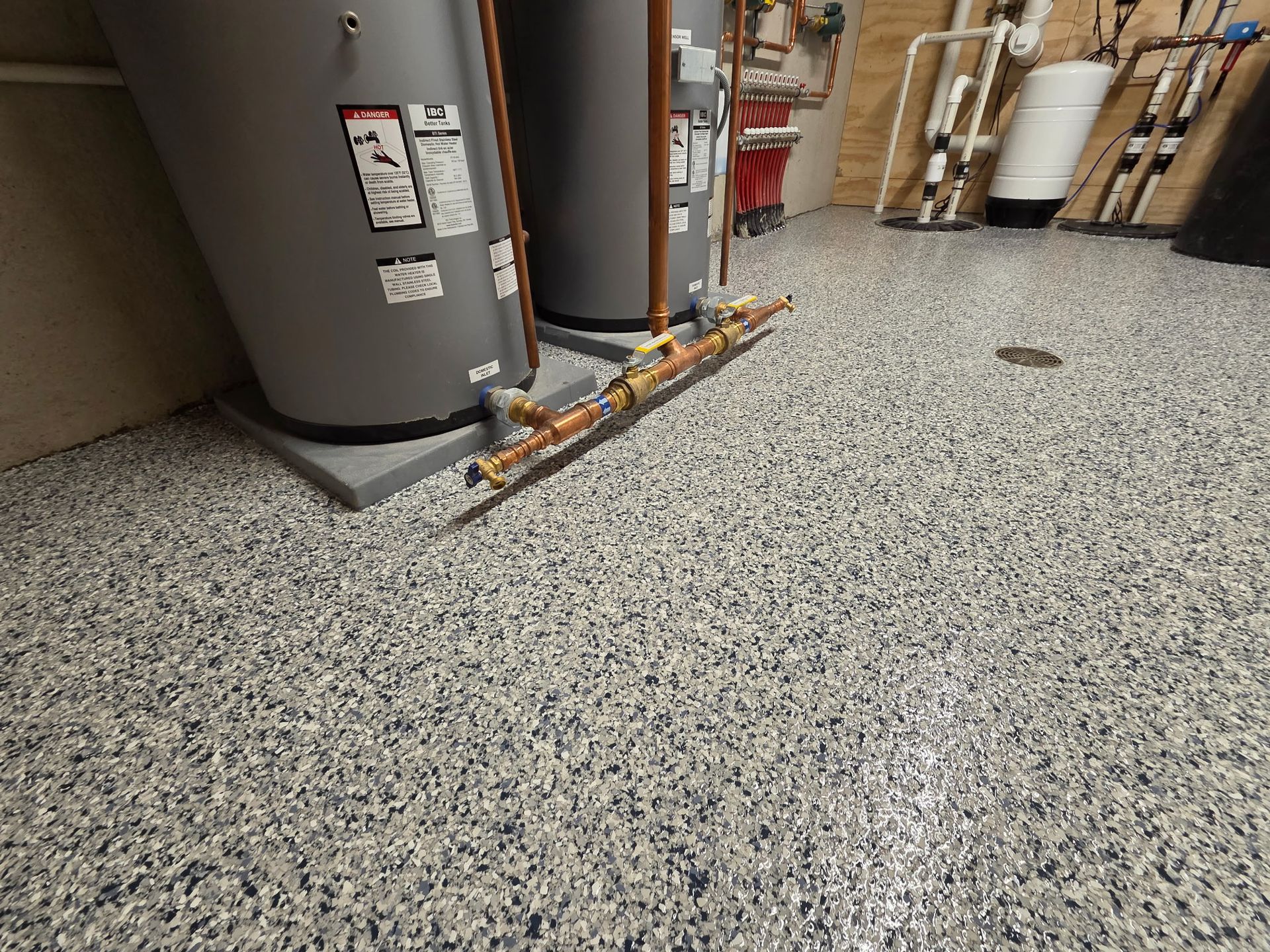 Utility room with water heater, exposed pipes, and speckled epoxy floor near a garage.