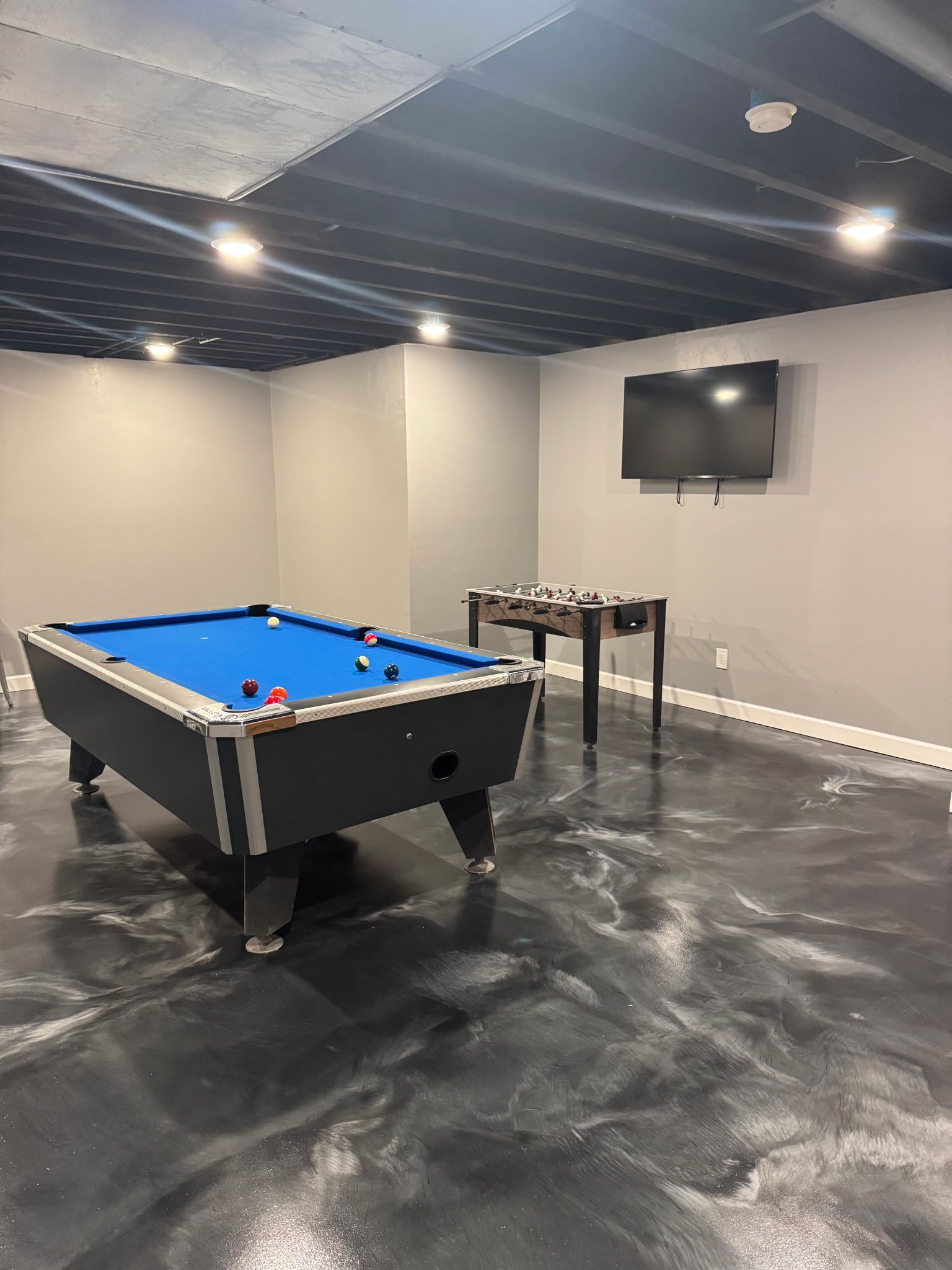 Modern game room with a blue pool table, TV, stools, and black polished floor