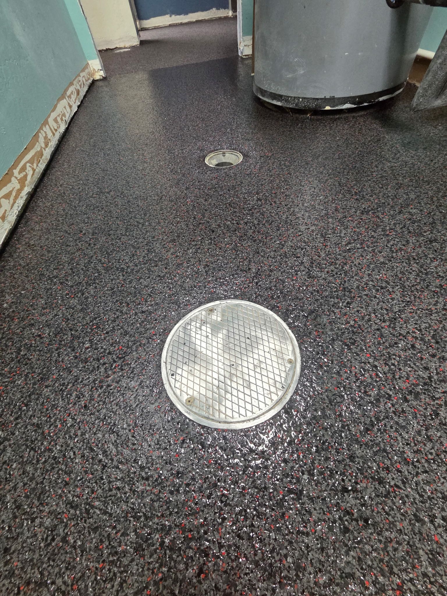 Black speckled floor with a round metal drain cover in a hallway.