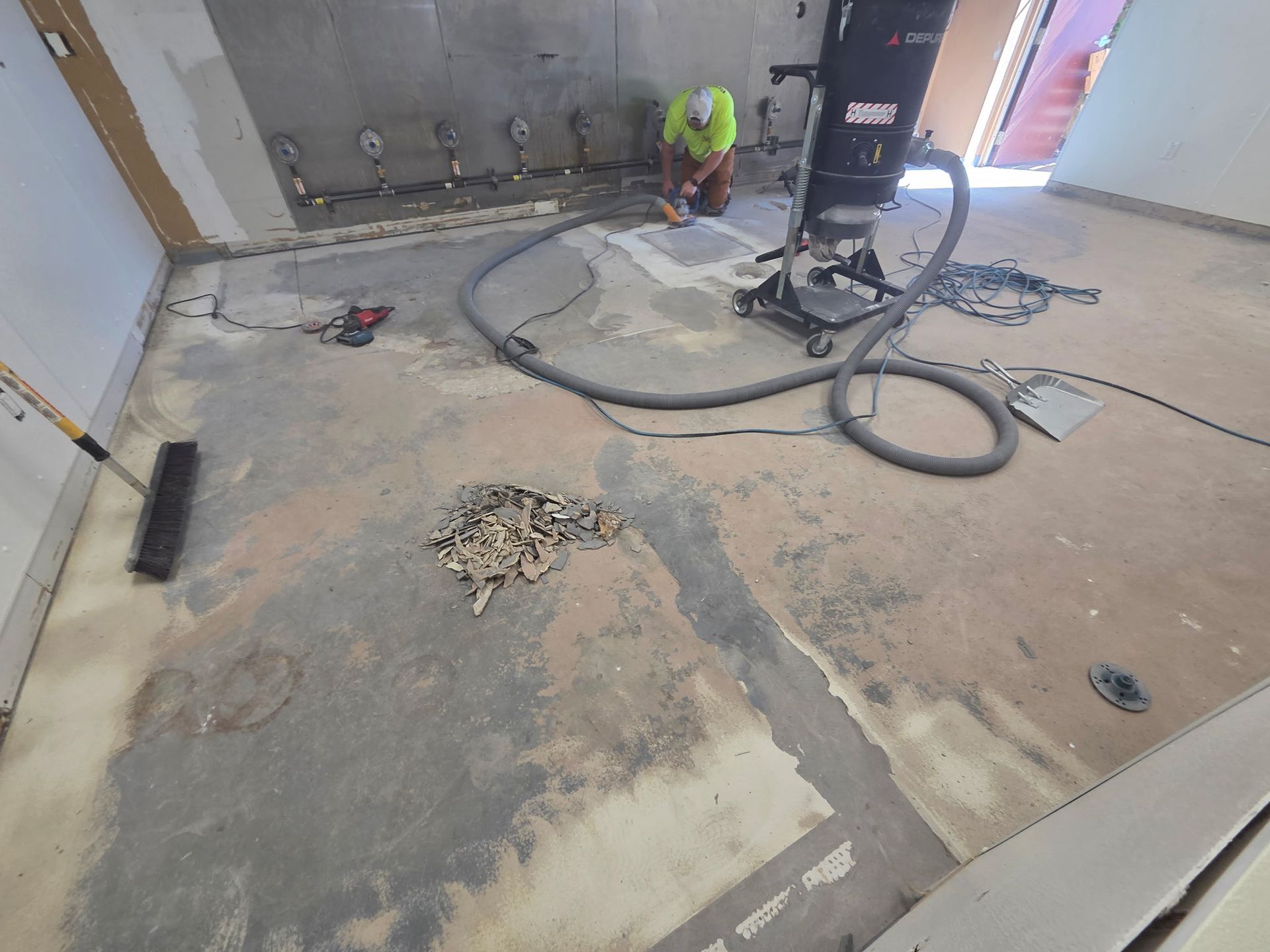 Worker grinding a concrete floor in a dusty room with tools, cables, and a large floor machine.