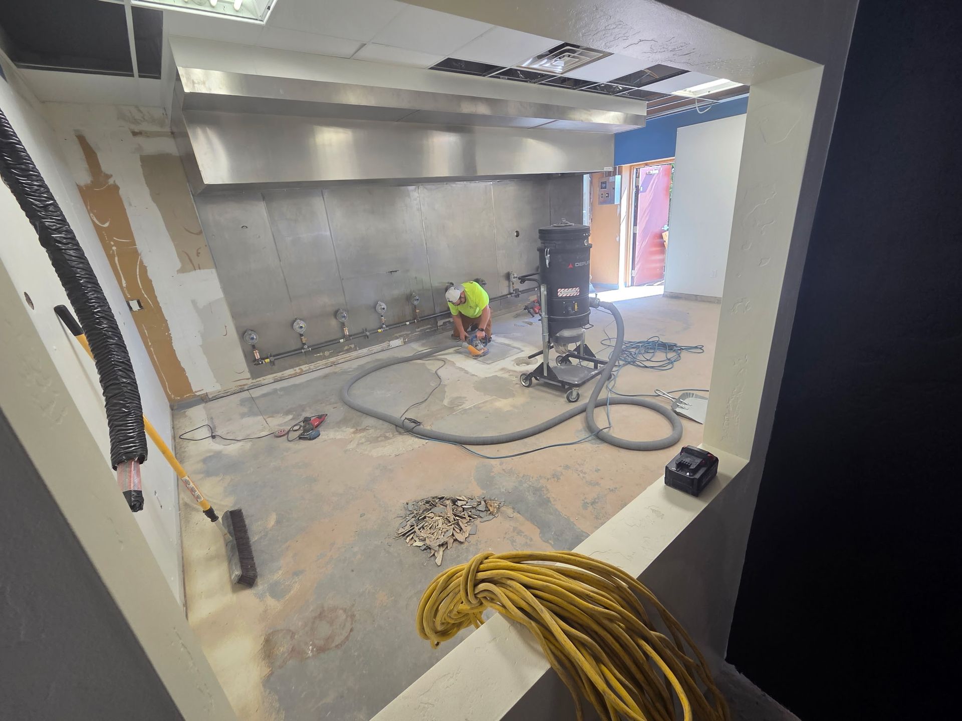 Worker in a fluorescent vest installing flooring in a large unfinished room with exposed wiring and tools.