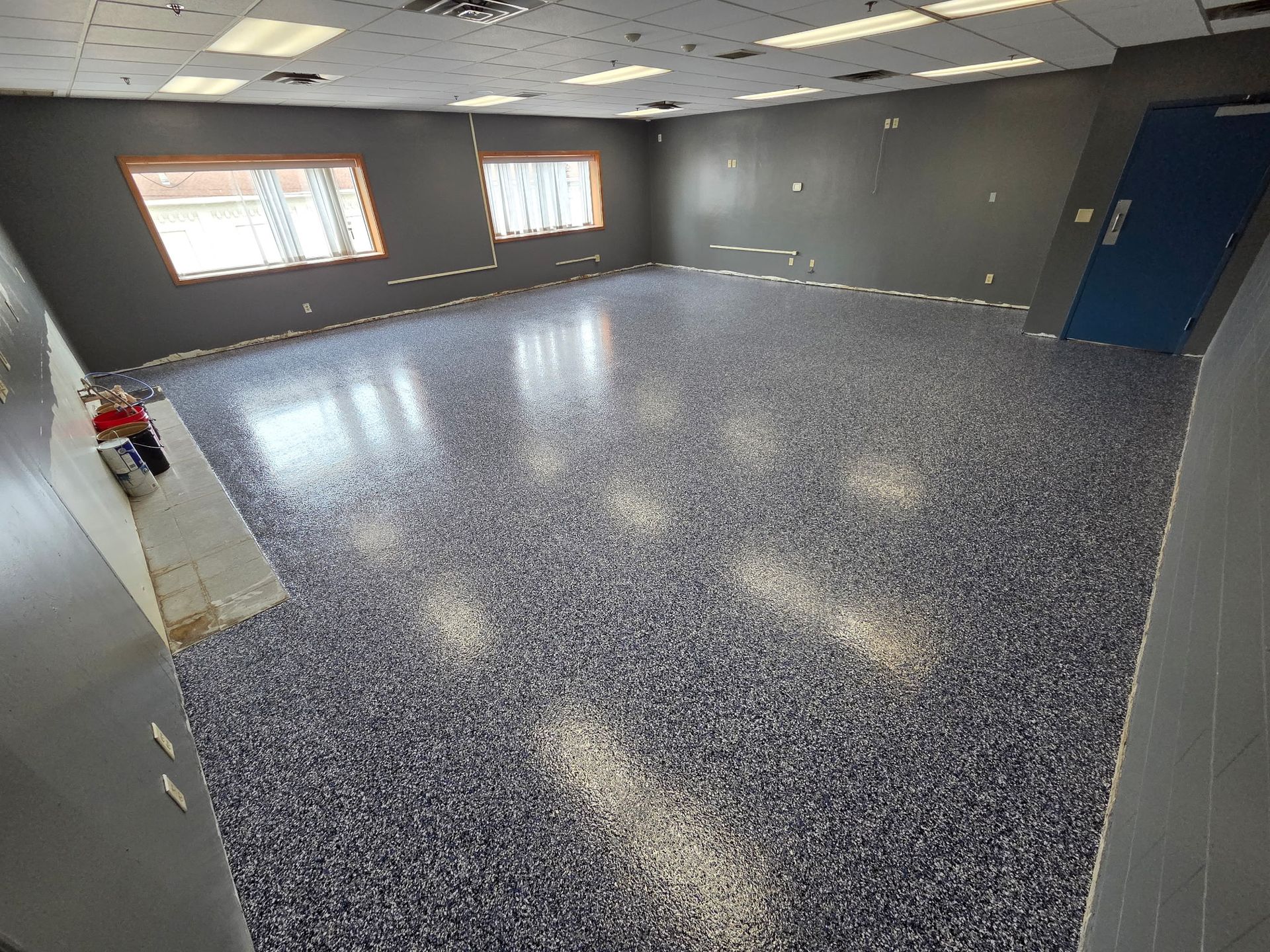 Empty gray room with speckled blue carpet, fluorescent lights, windows, and a blue door