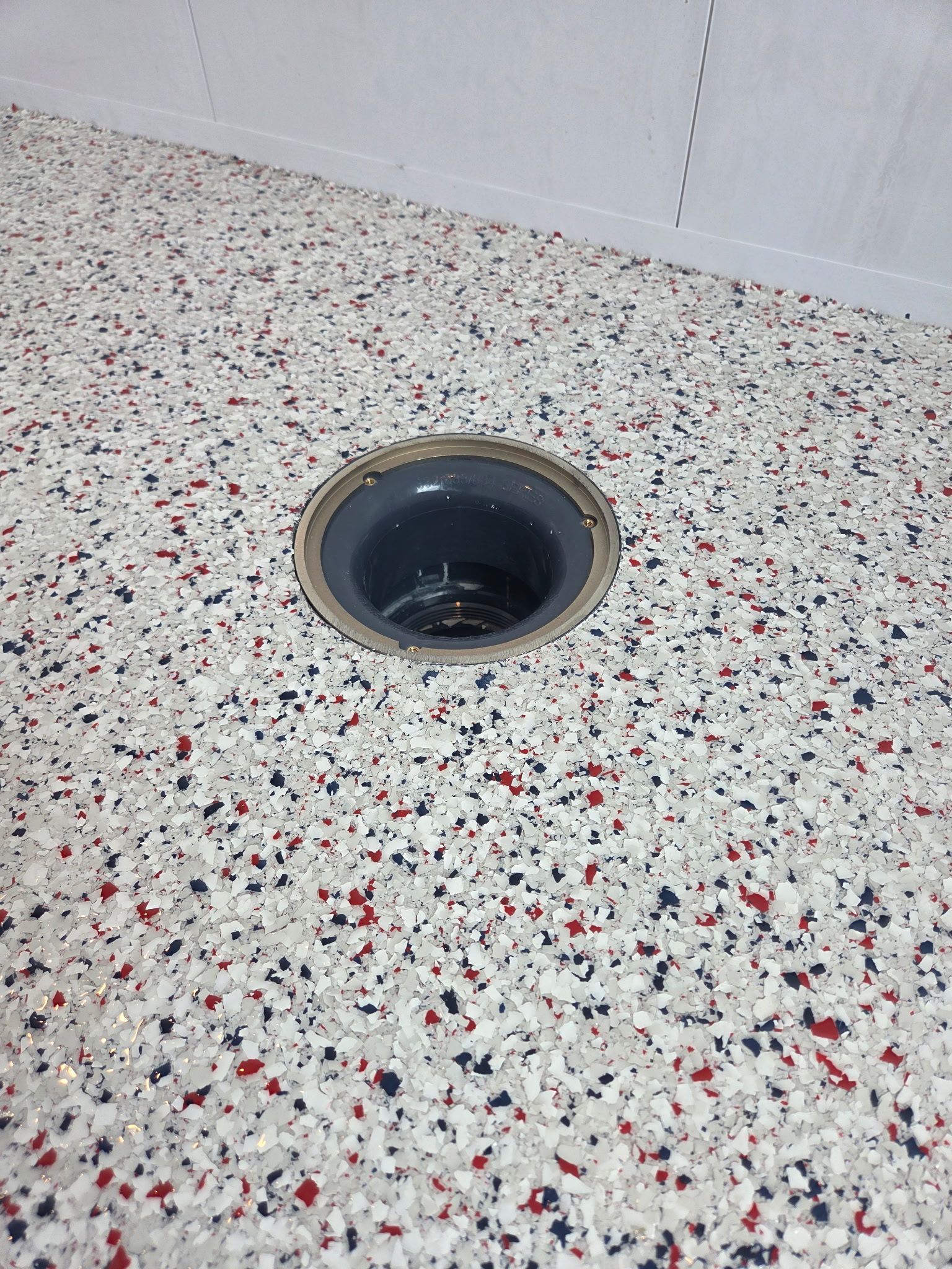 A floor drain set in a speckled terrazzo floor near a tiled wall.