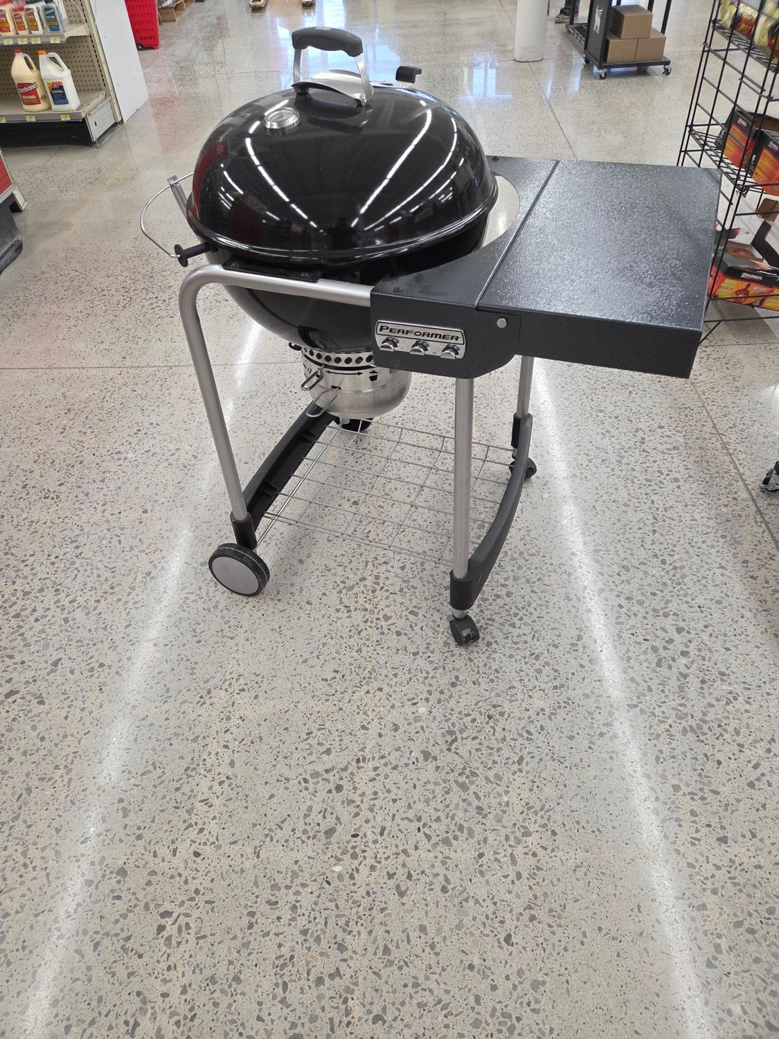 A black charcoal kettle grill with a side table on a wheeled metal frame, standing on a light-colored tiled floor