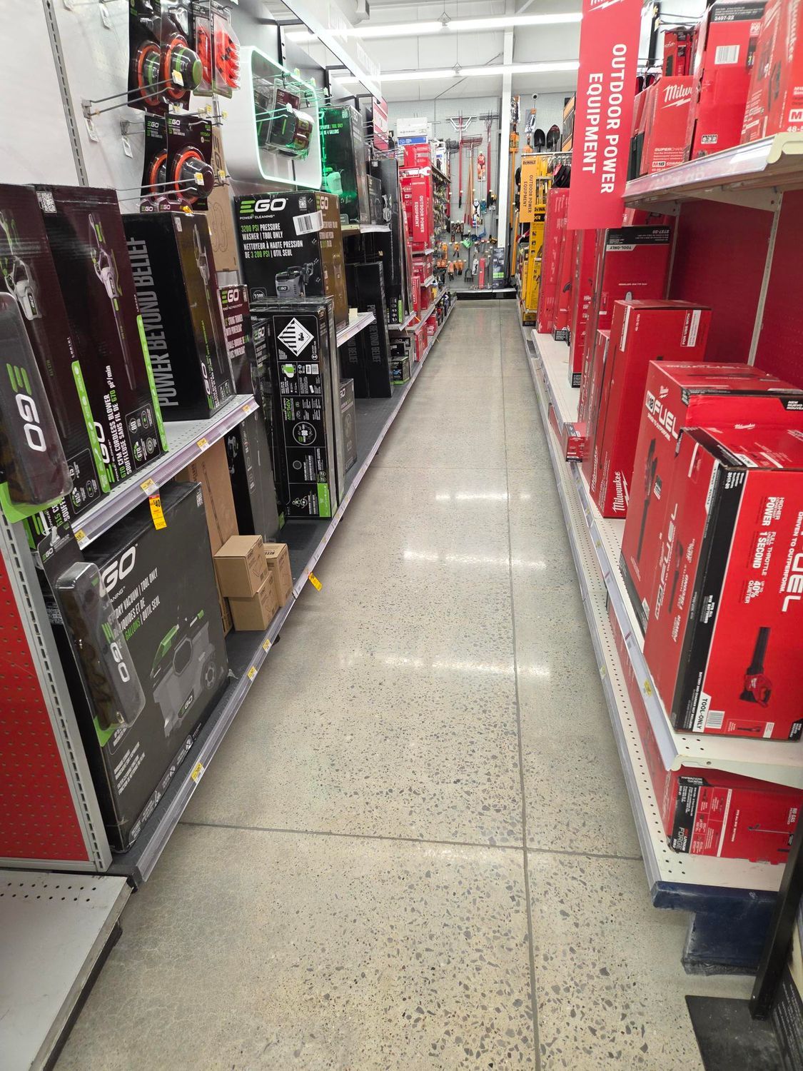 A retail aisle lined with shelves filled with packaged power equipment and red boxes, leading toward a bright back wall