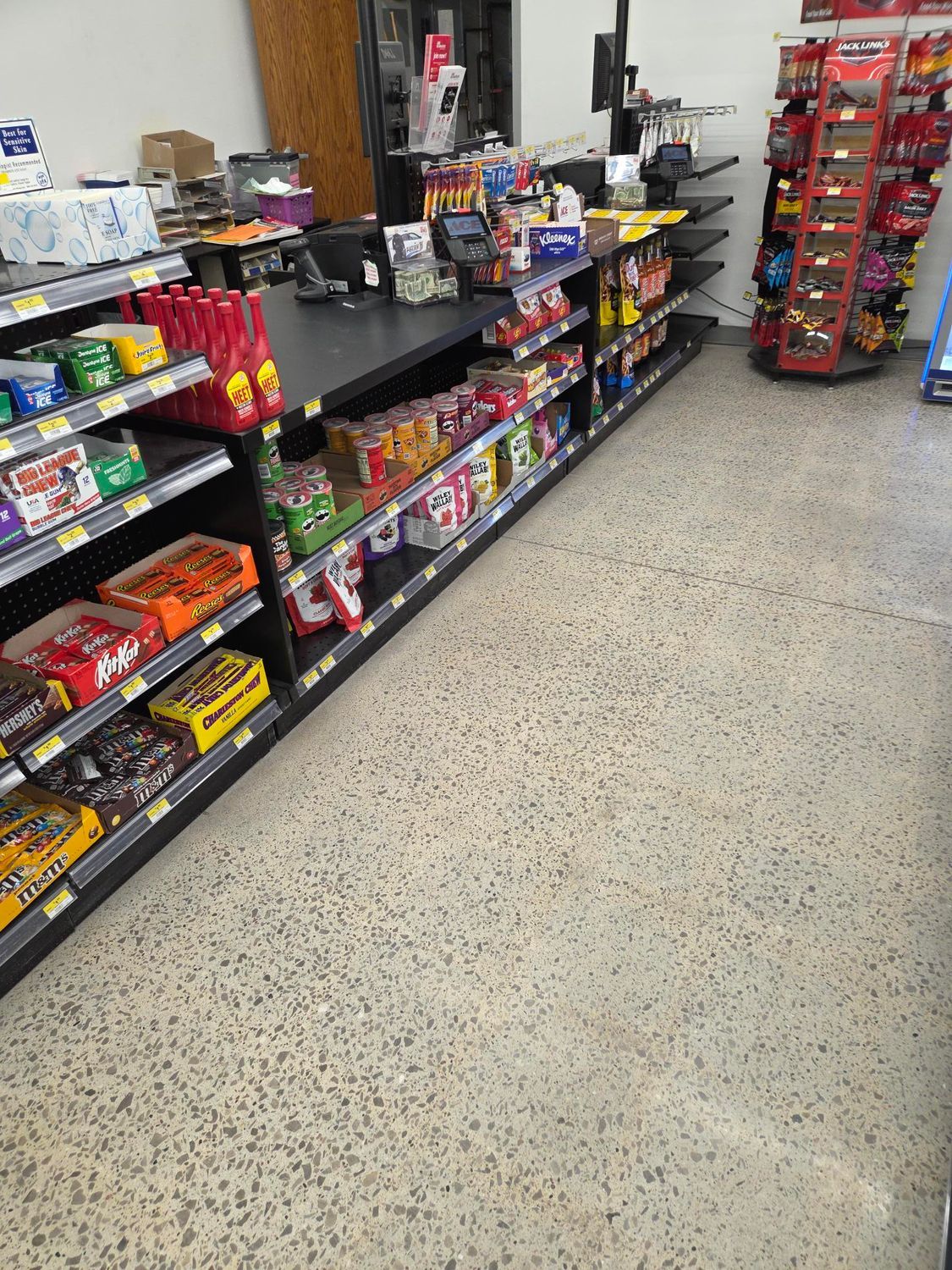 Convenience store aisle with shelves of snacks, candy, and drinks beside a speckled floor.