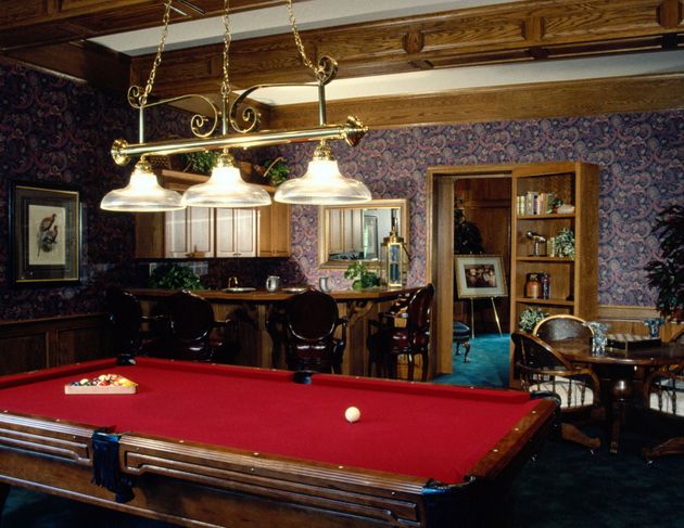 A billiards room featuring a red felt pool table, a wooden bar with stools, and patterned wallpaper.