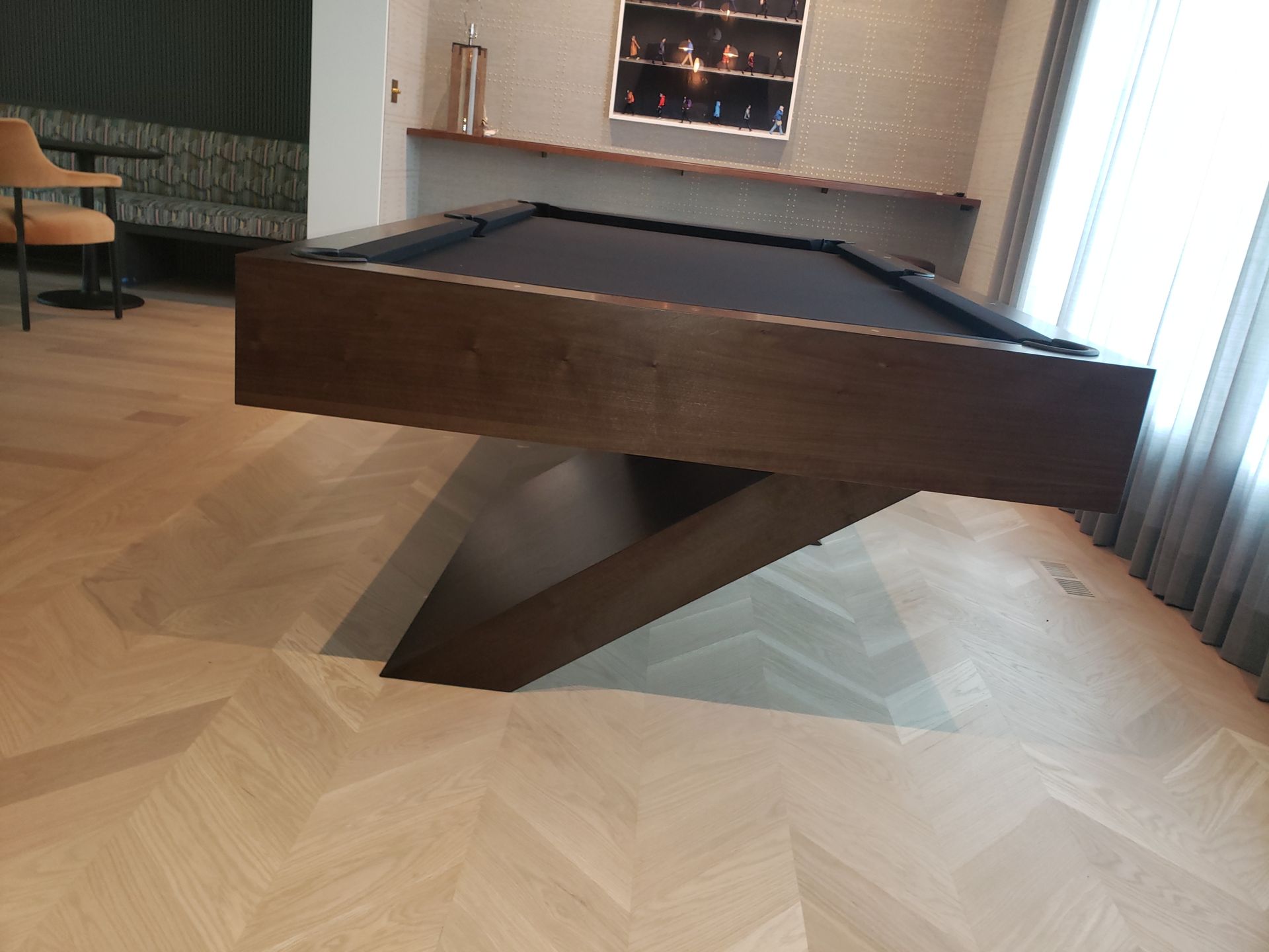 A modern pool table with a dark felt top and unique angular pedestal base sits on a light herringbone wood floor.