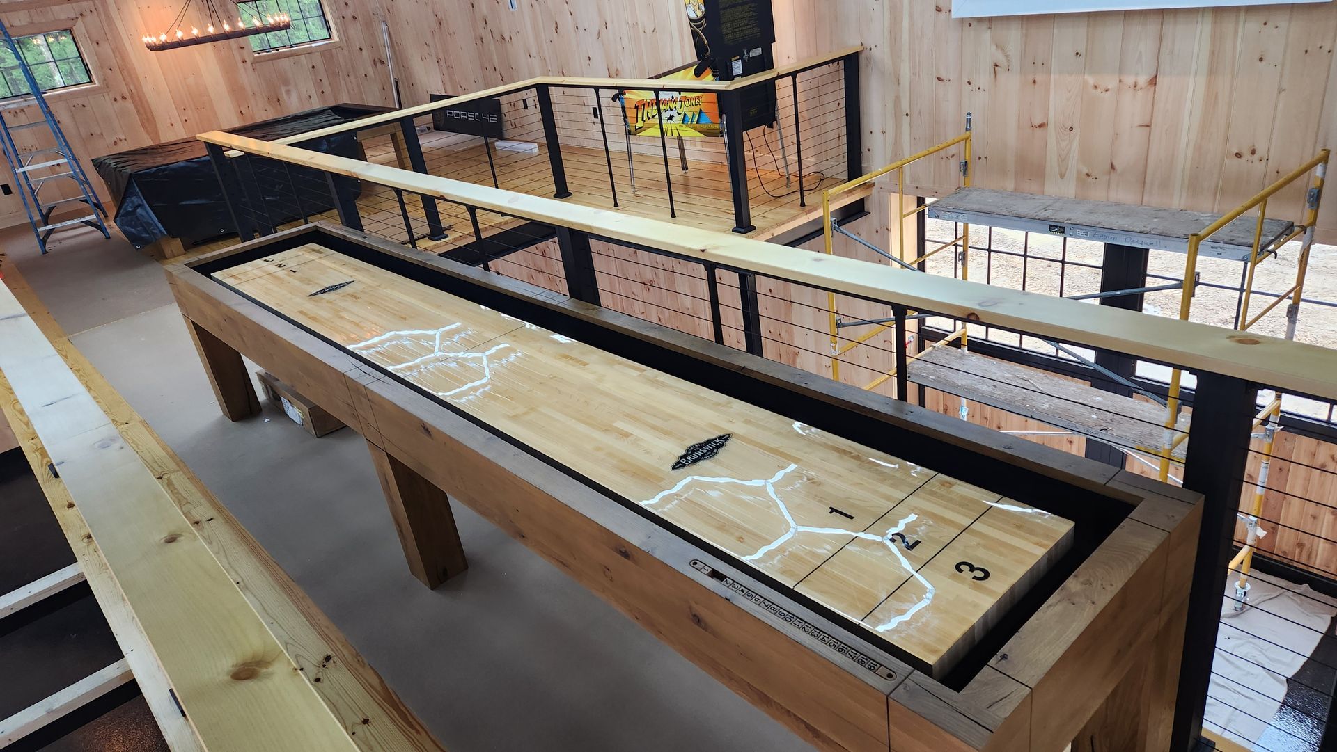 A wooden shuffleboard table inside a rustic game room with wood-paneled walls, metal railings, and additional games.