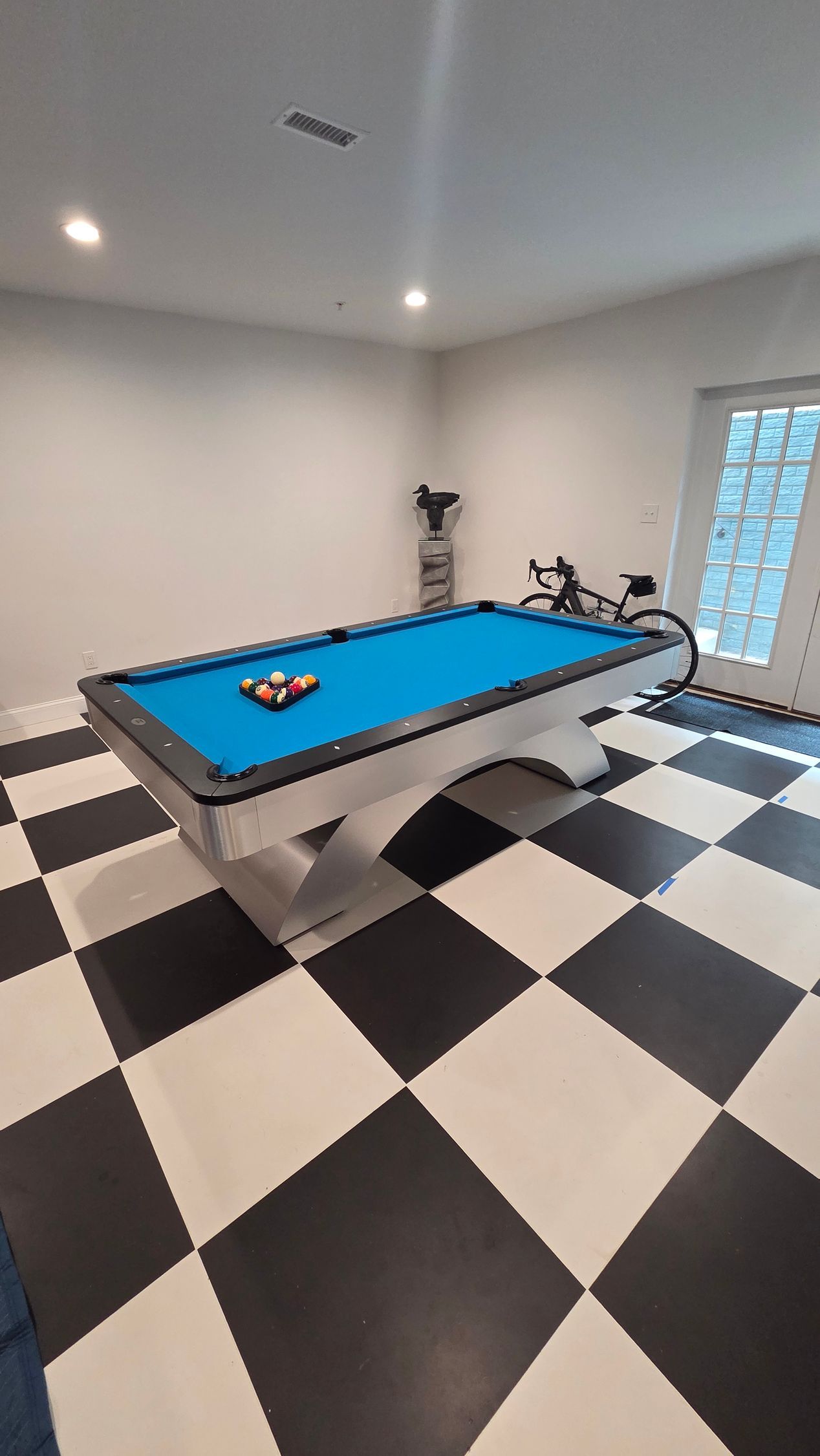 A blue pool table sits in the center of a room with a black-and-white checkered floor and a bicycle by a glass door.