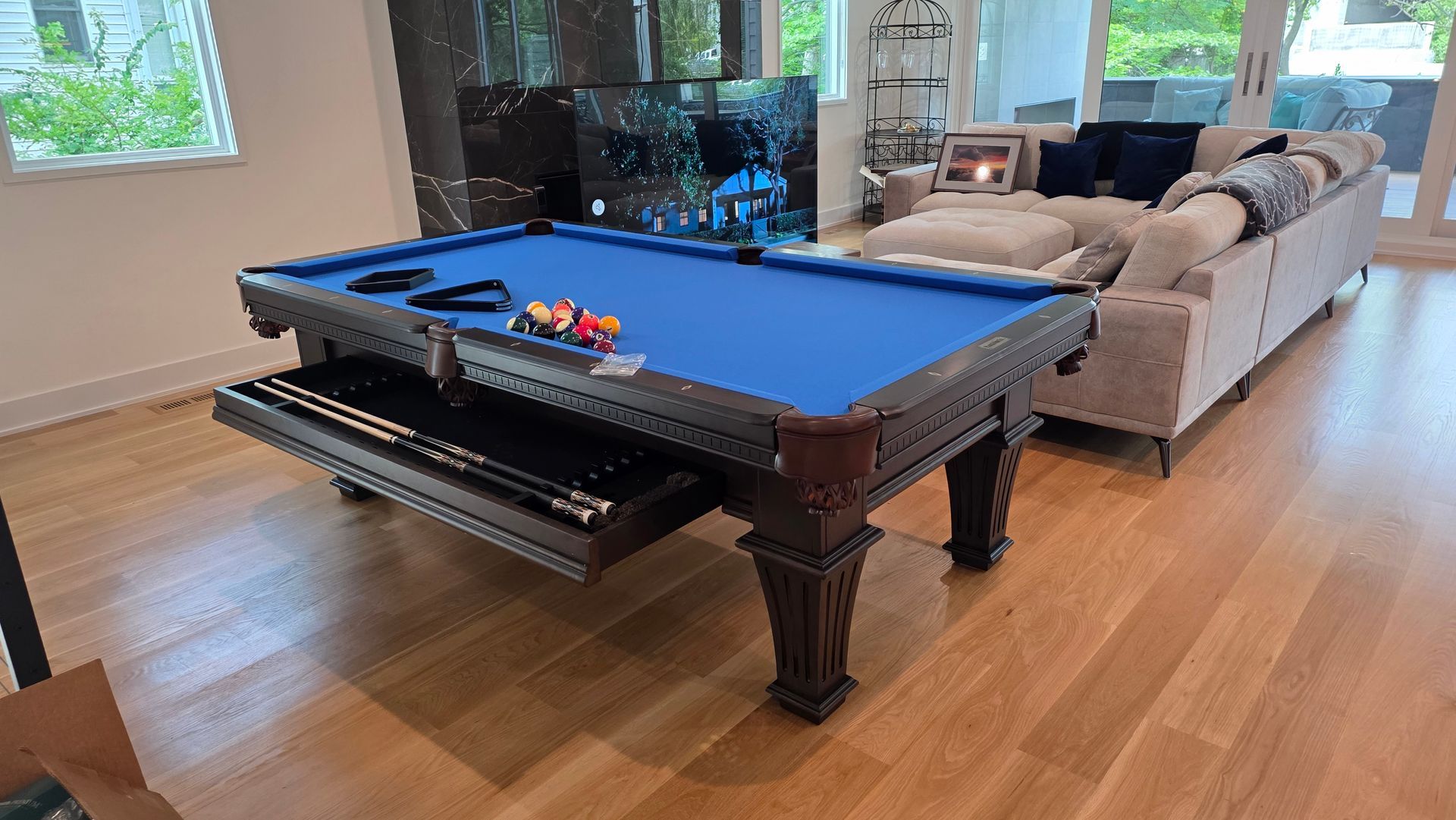 A blue pool table with an open storage drawer containing cues sits on a light hardwood floor in a living room.