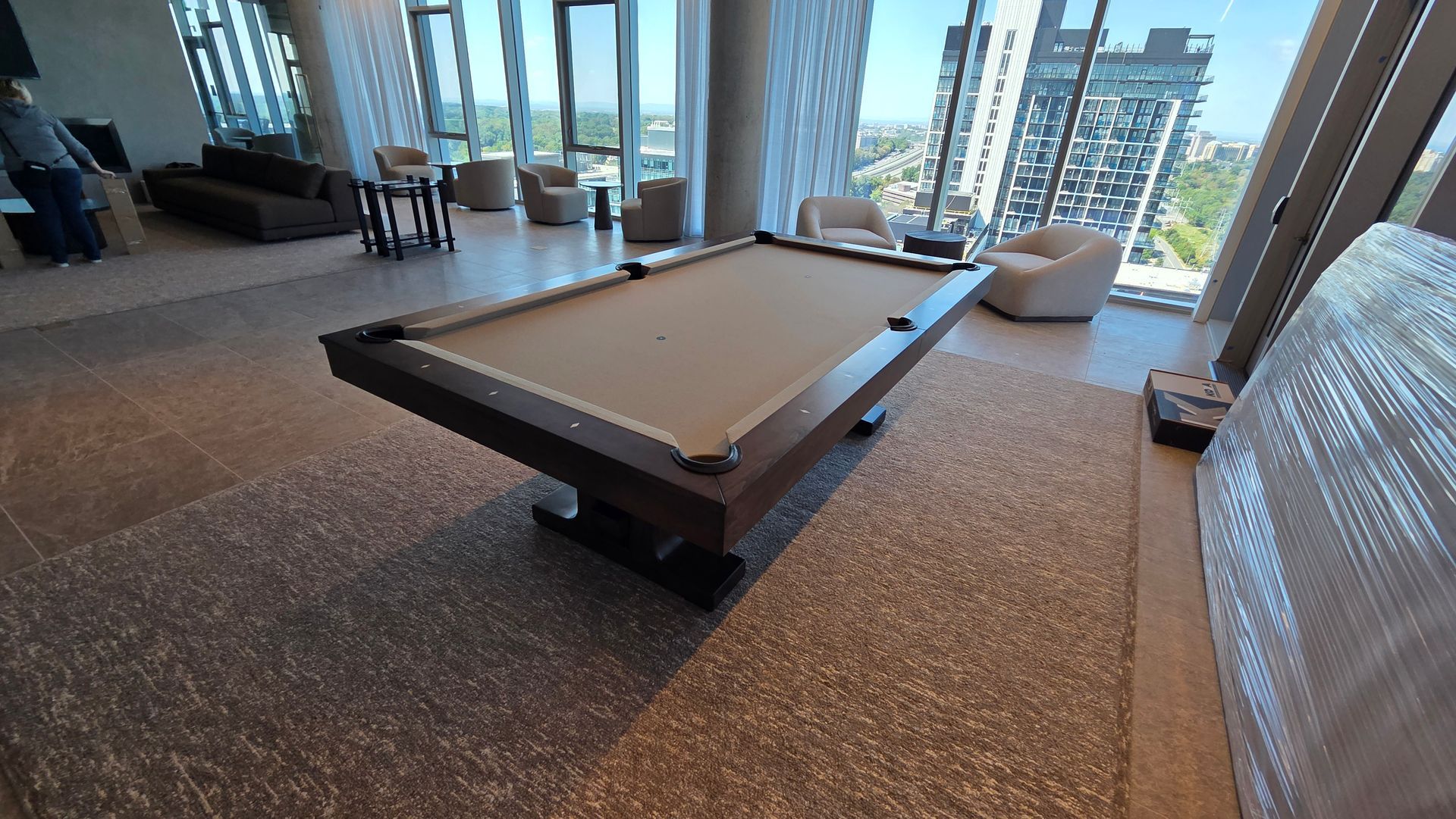 A lounge area with a tan-felt pool table in the foreground, surrounded by armchairs, sofas, and floor-to-ceiling windows.