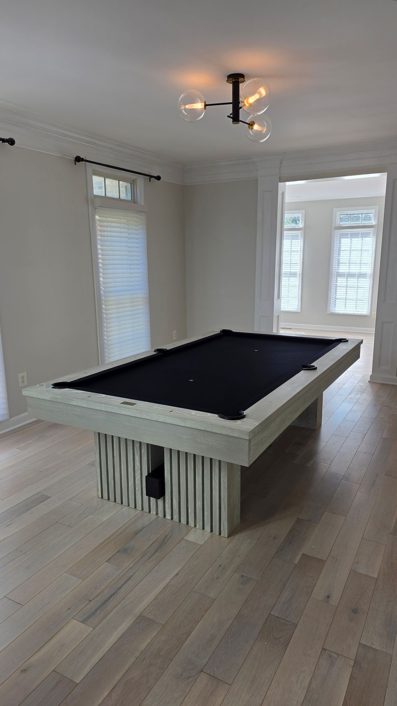 A modern light-wood pool table with a black felt top sits in a bright, minimalist room with wood floors.