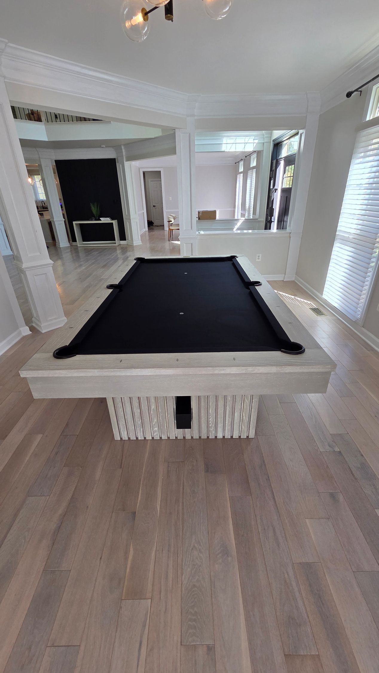 A black pool table with a light, textured base sits in a bright, spacious home with hardwood floors and white trim.