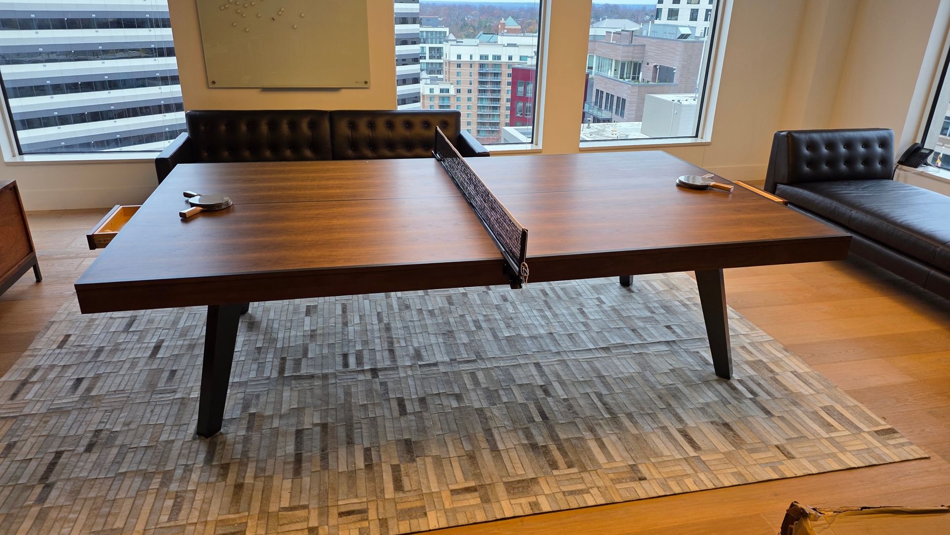 A dark wood ping pong table sits on a patterned rug in a bright, modern office with a view of city buildings.