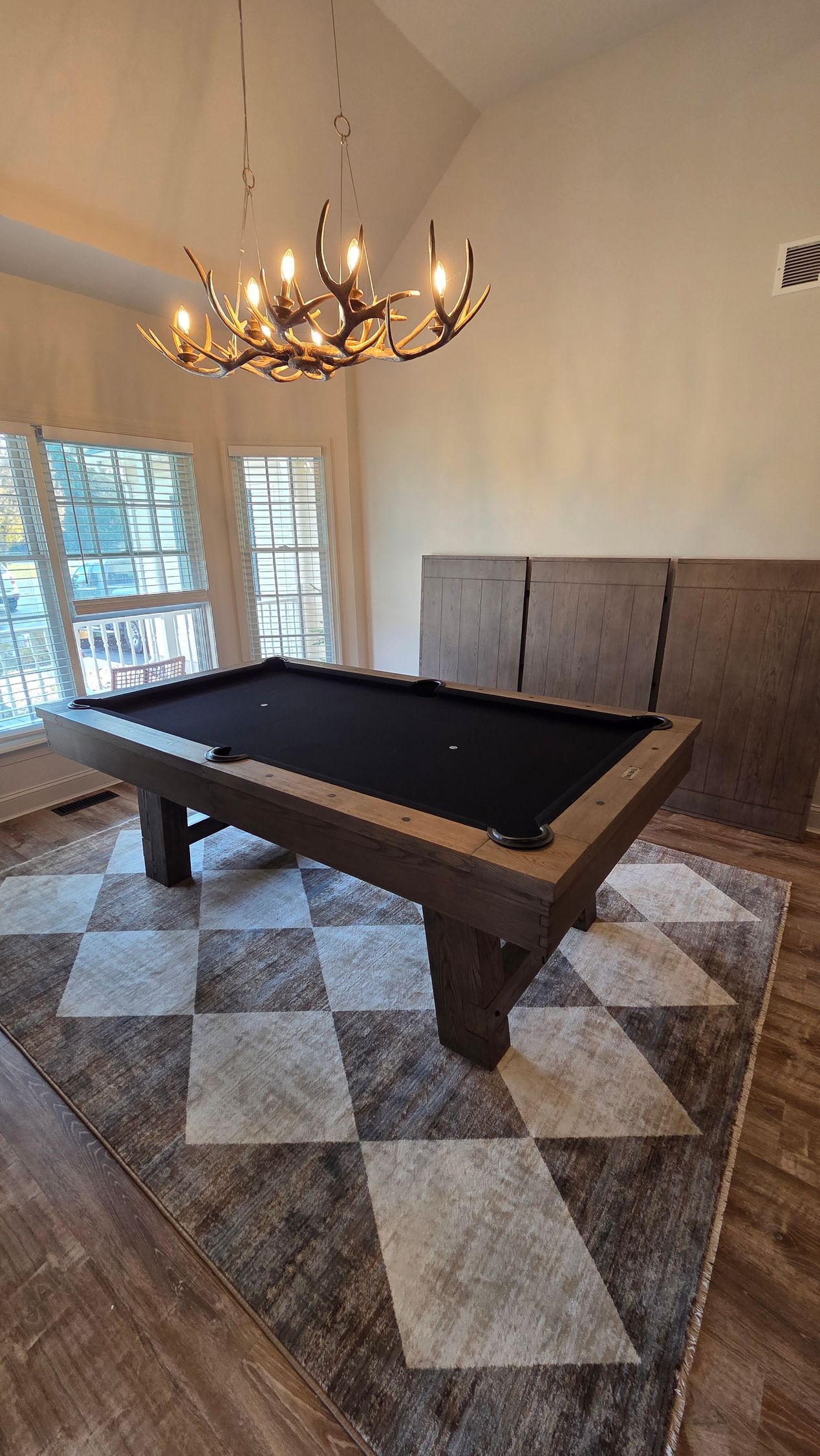A pool table with a black felt surface sits on a gray and white diamond-patterned rug beneath an antler chandelier.