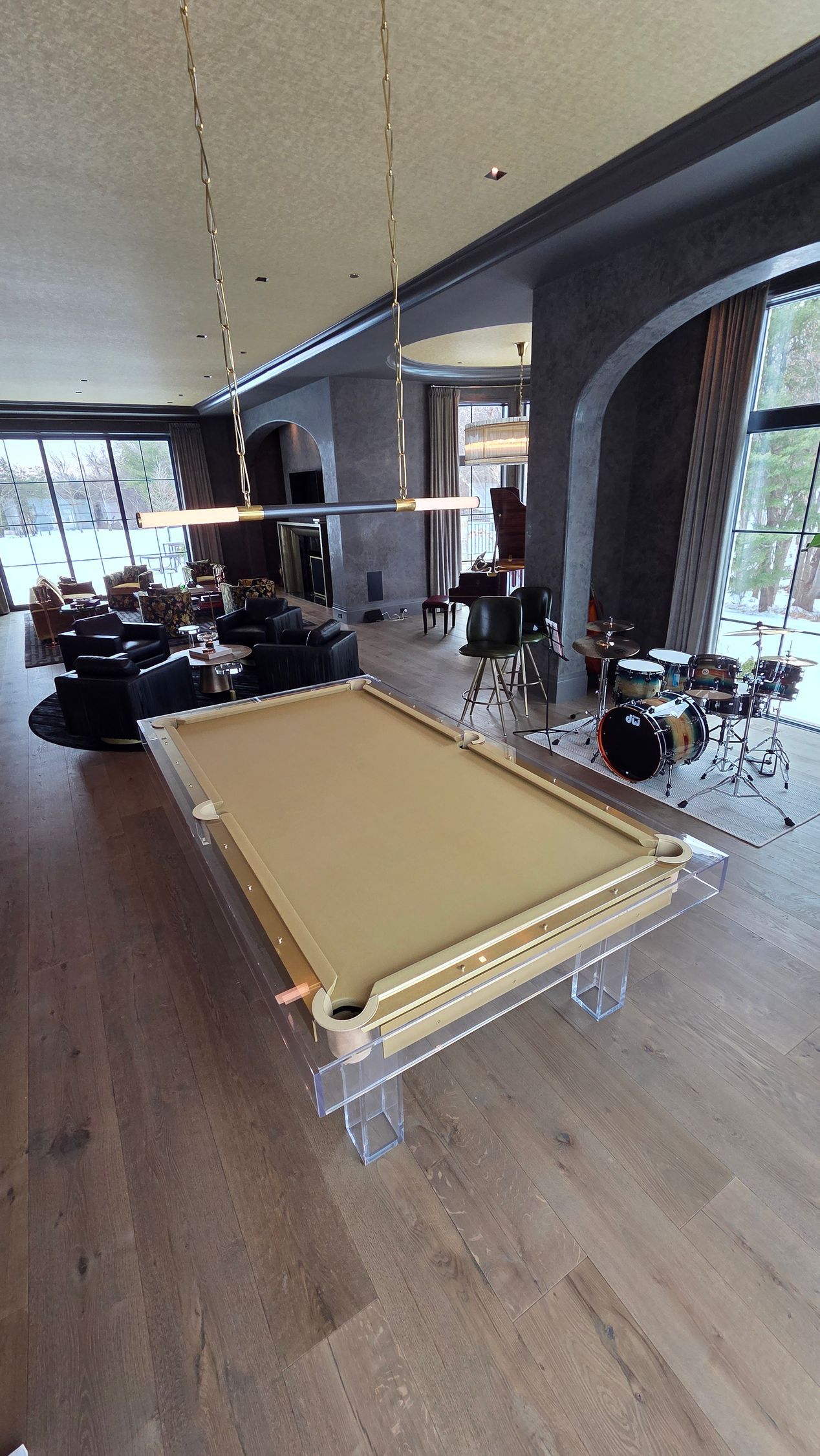A clear, acrylic-frame pool table with a tan felt top stands in a spacious, modern room with a drum kit in the background.