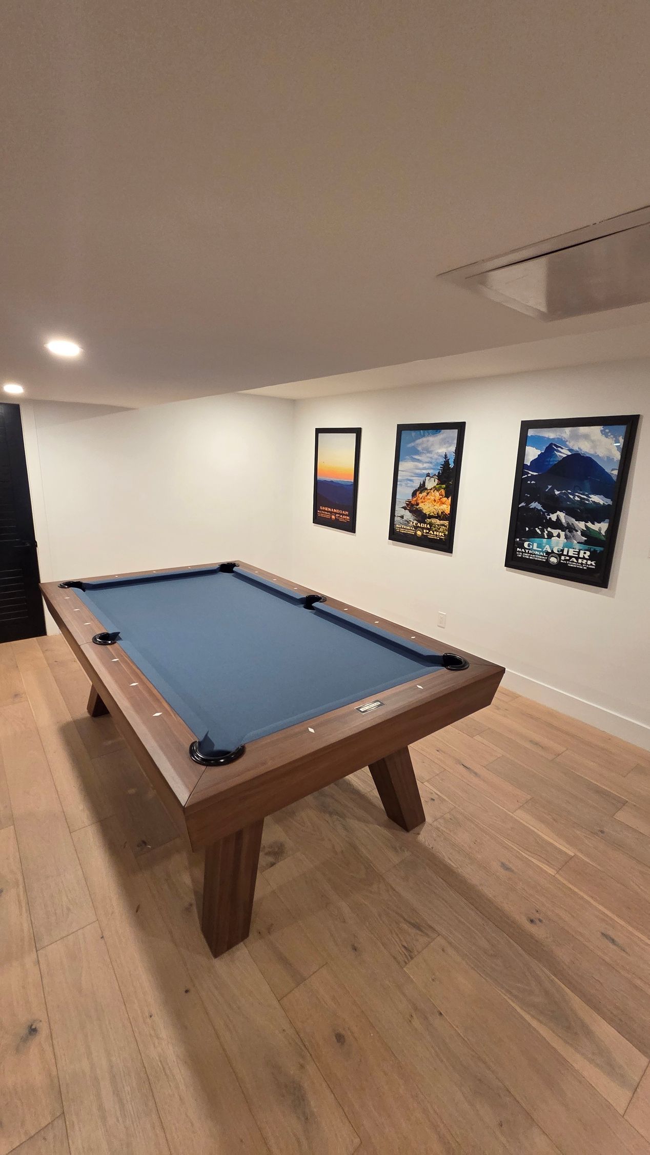 A pool table with blue felt sits in a modern, light-flooded room with three framed landscape posters on the wall.