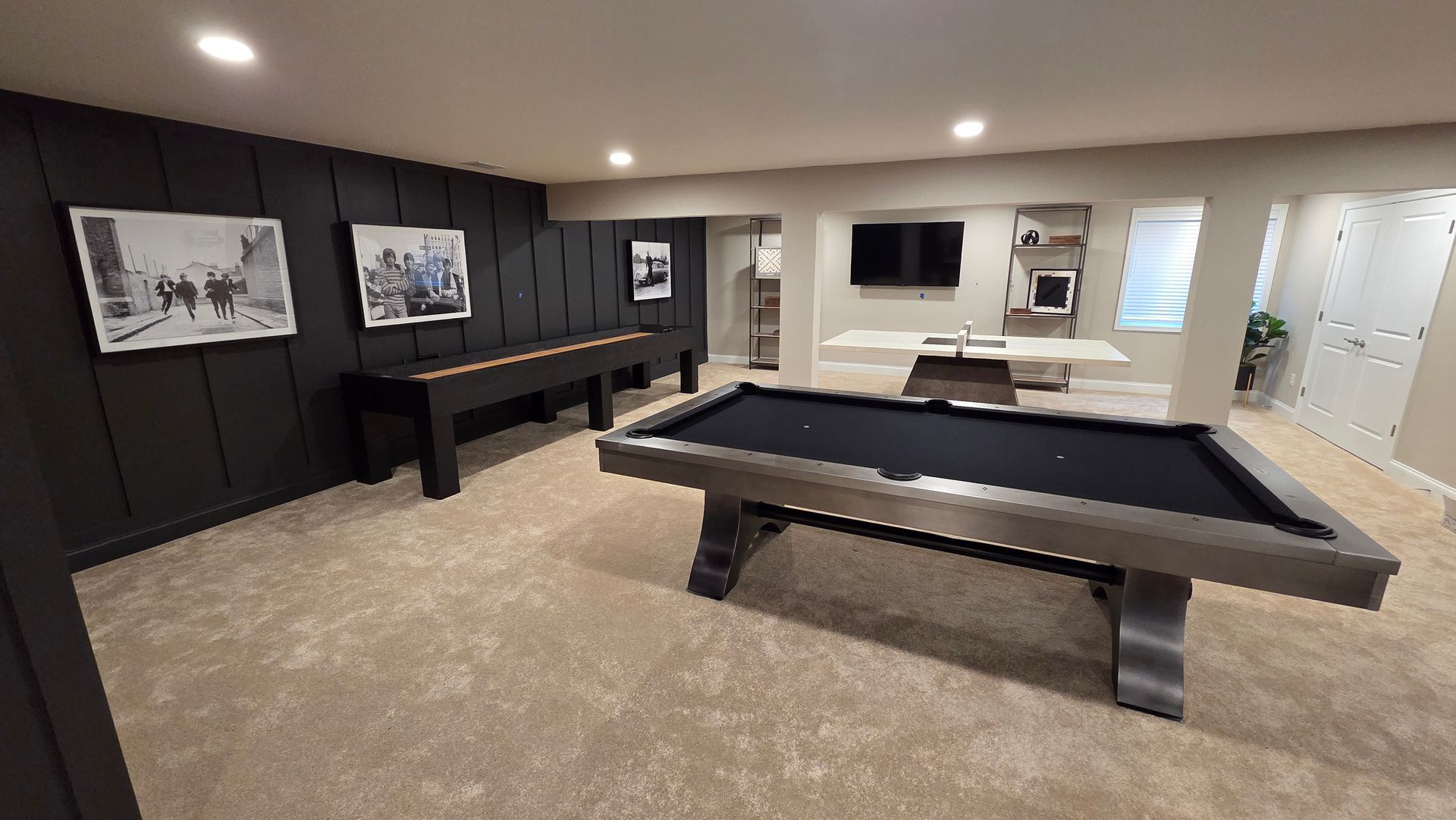 A basement game room featuring a pool table, shuffleboard table, dark wood-paneled wall with art, and television.