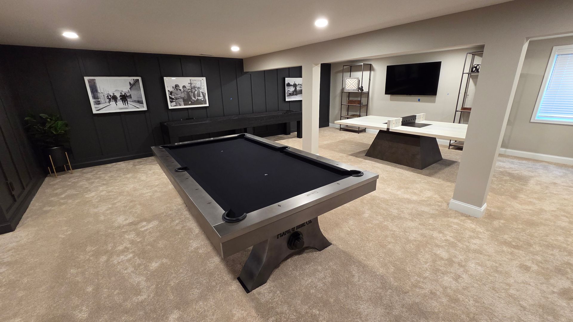 A basement game room featuring a black pool table, a ping pong table, and framed artwork on a dark accent wall.
