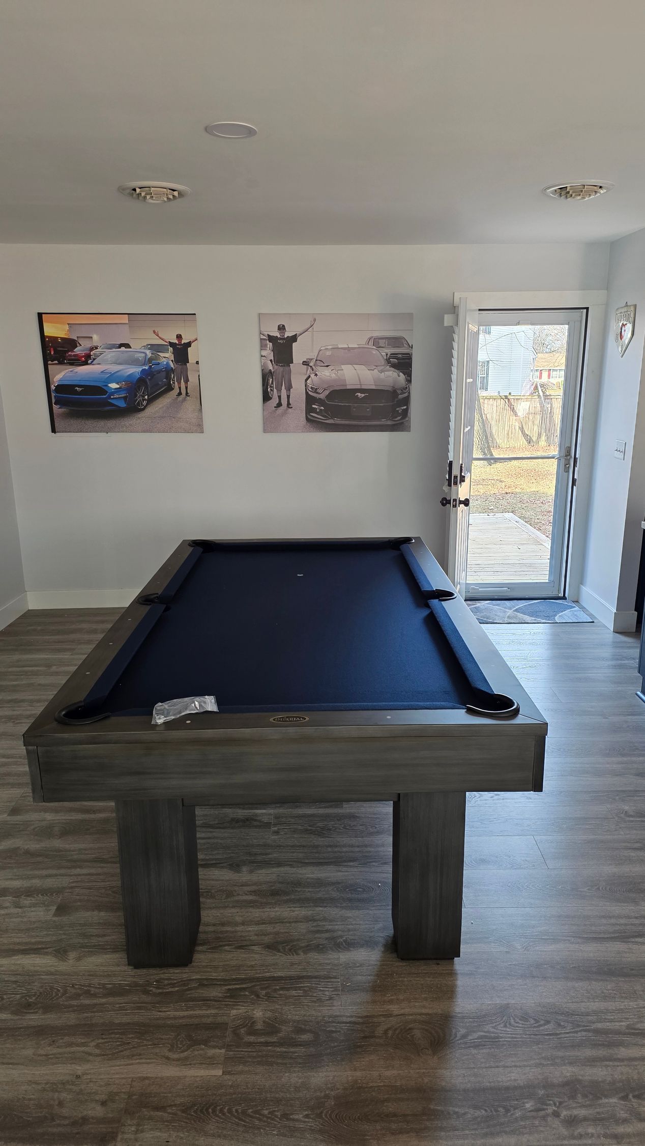 A pool table with a dark blue cloth stands in a room with light wood floors, two car posters on the wall, and a doorway.