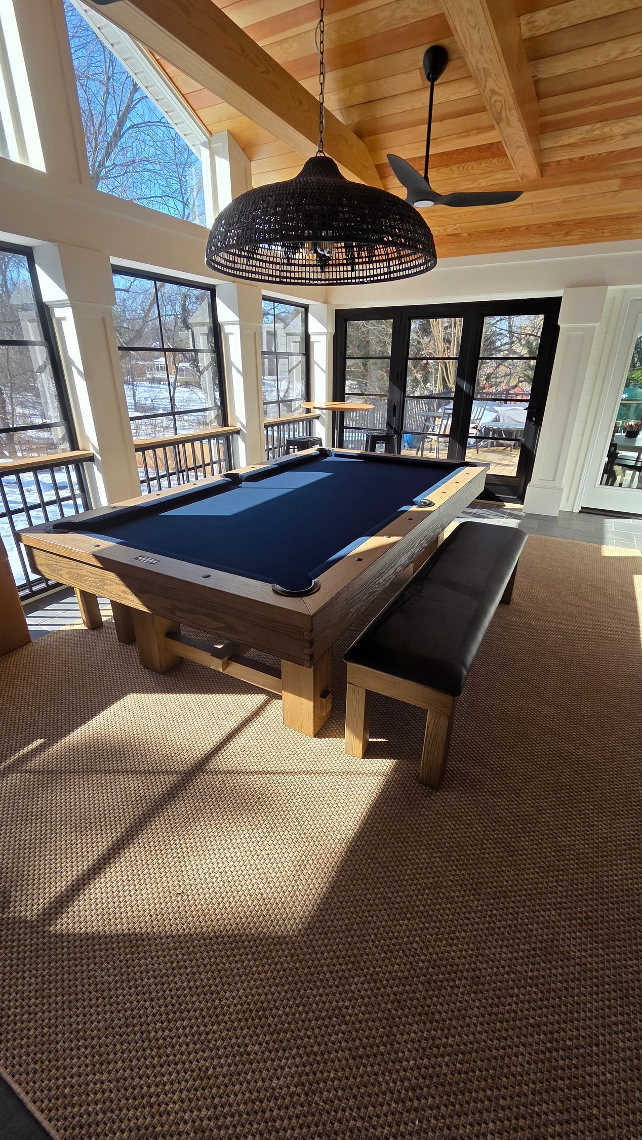 A billiards table with a dark blue felt surface and a matching bench seat in a sunlit, wood-paneled indoor porch.