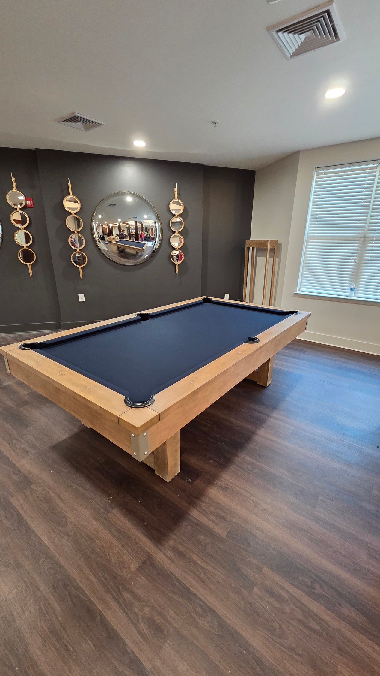 A room featuring a pool table with navy blue felt and light wood framing, centered before a wall with hanging decor.