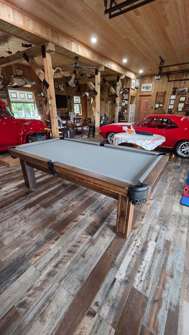 A billiard table with a gray felt surface centered in a rustic garage featuring wooden beams and two red classic cars.
